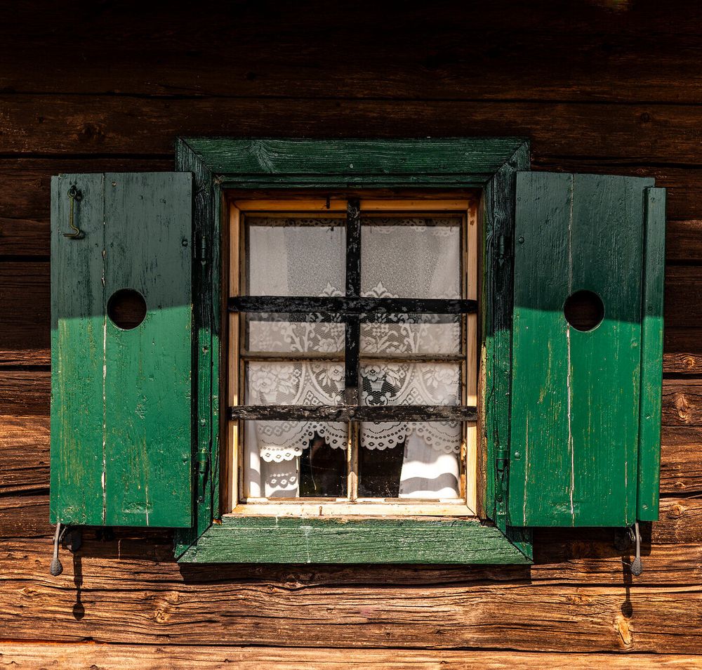 Fenster einer urigen Almhütte mit Fensterläden und Häkelgardine.