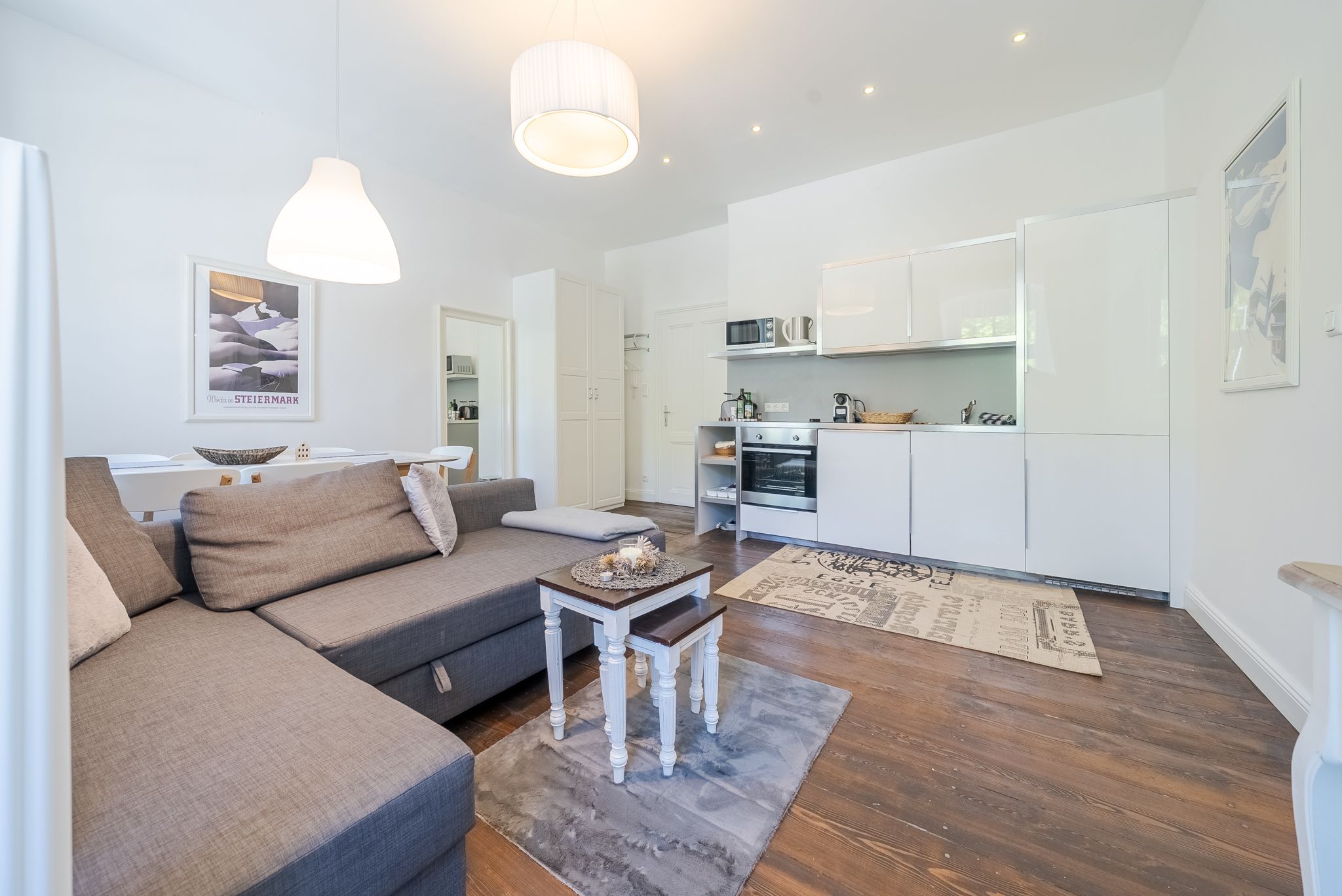Modern eingerichtetes Wohnzimmer mit grauem Sofa, weißer Küche und Holzboden.