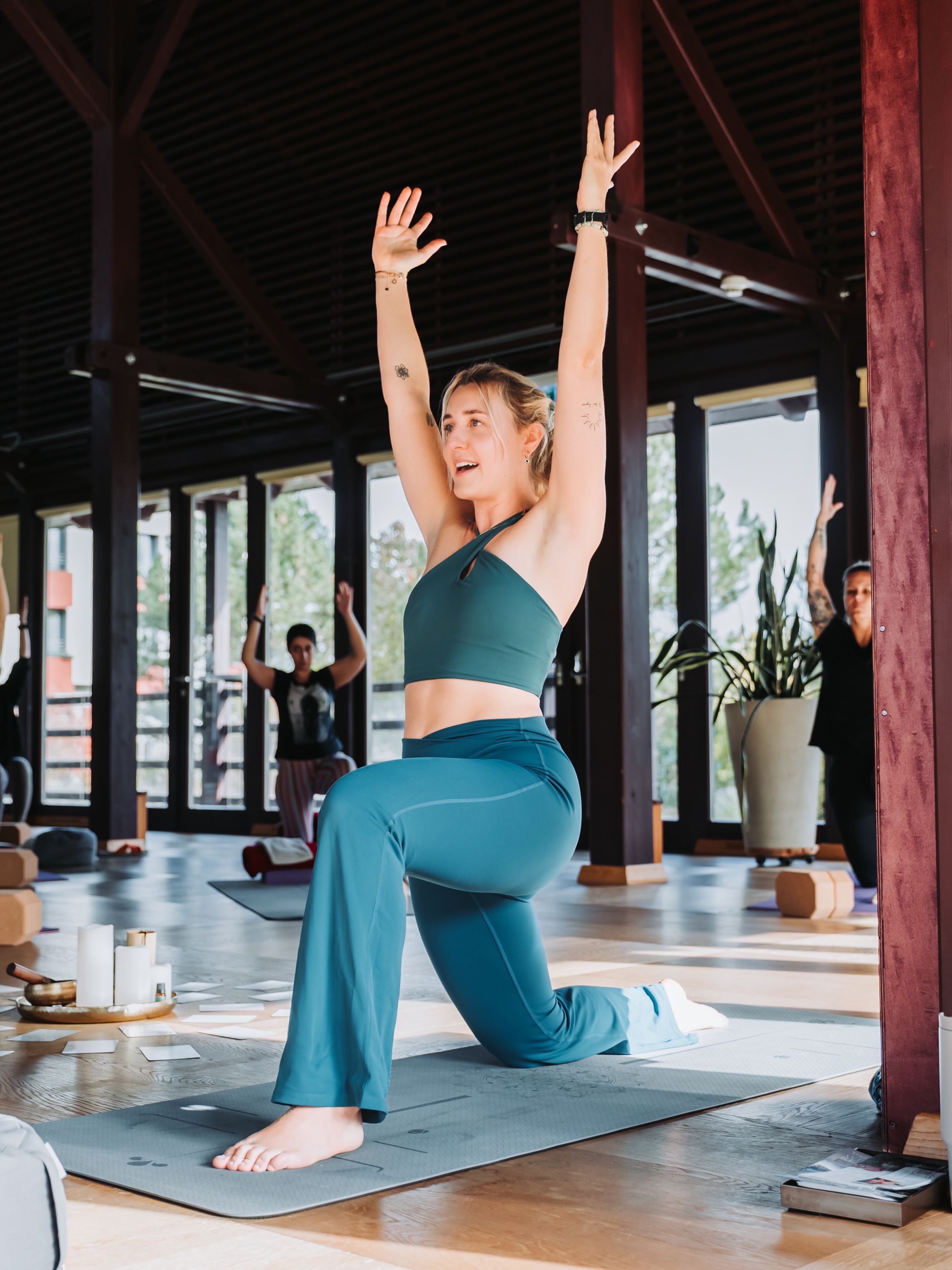 Eine Frau in blauer Sportkleidung macht eine Yoga-Übung in einem Raum mit Holzboden und großen Fenstern.
