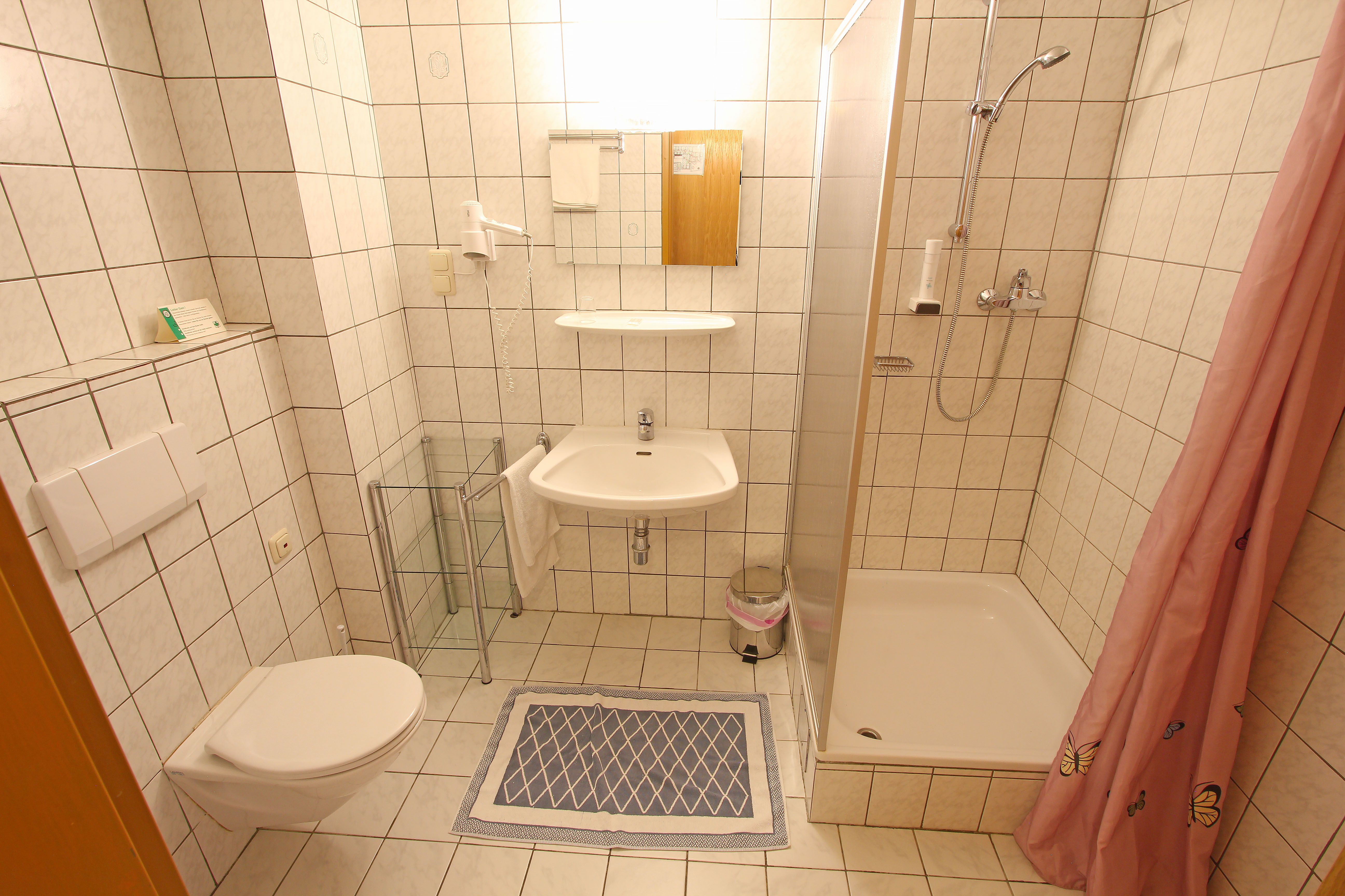 Kleines Badezimmer mit Dusche, Waschbecken und Toilette, gefliest in Weiß.