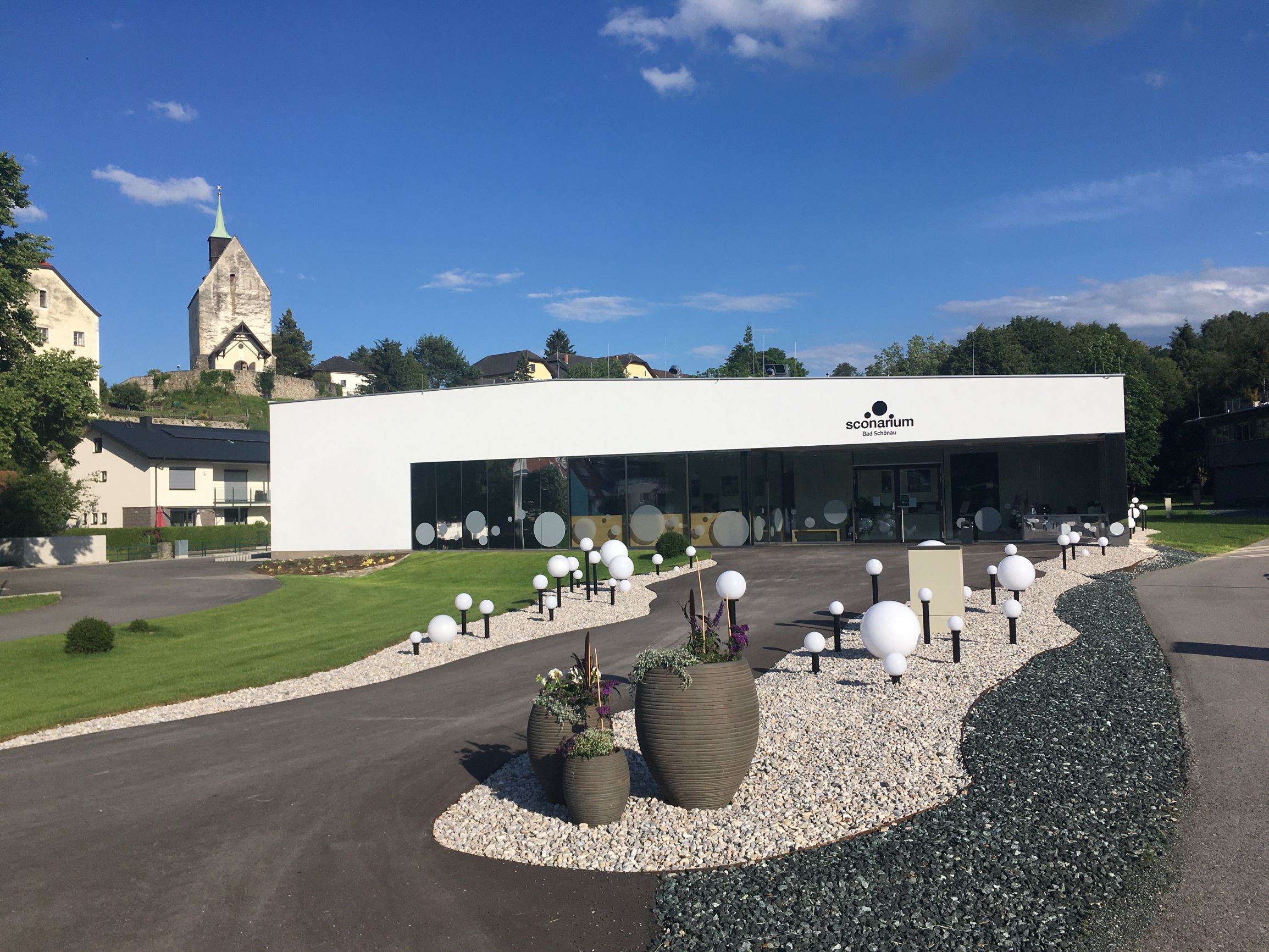 Modernes Gebäude des Sconariums mit Garten und Kirche im Hintergrund.