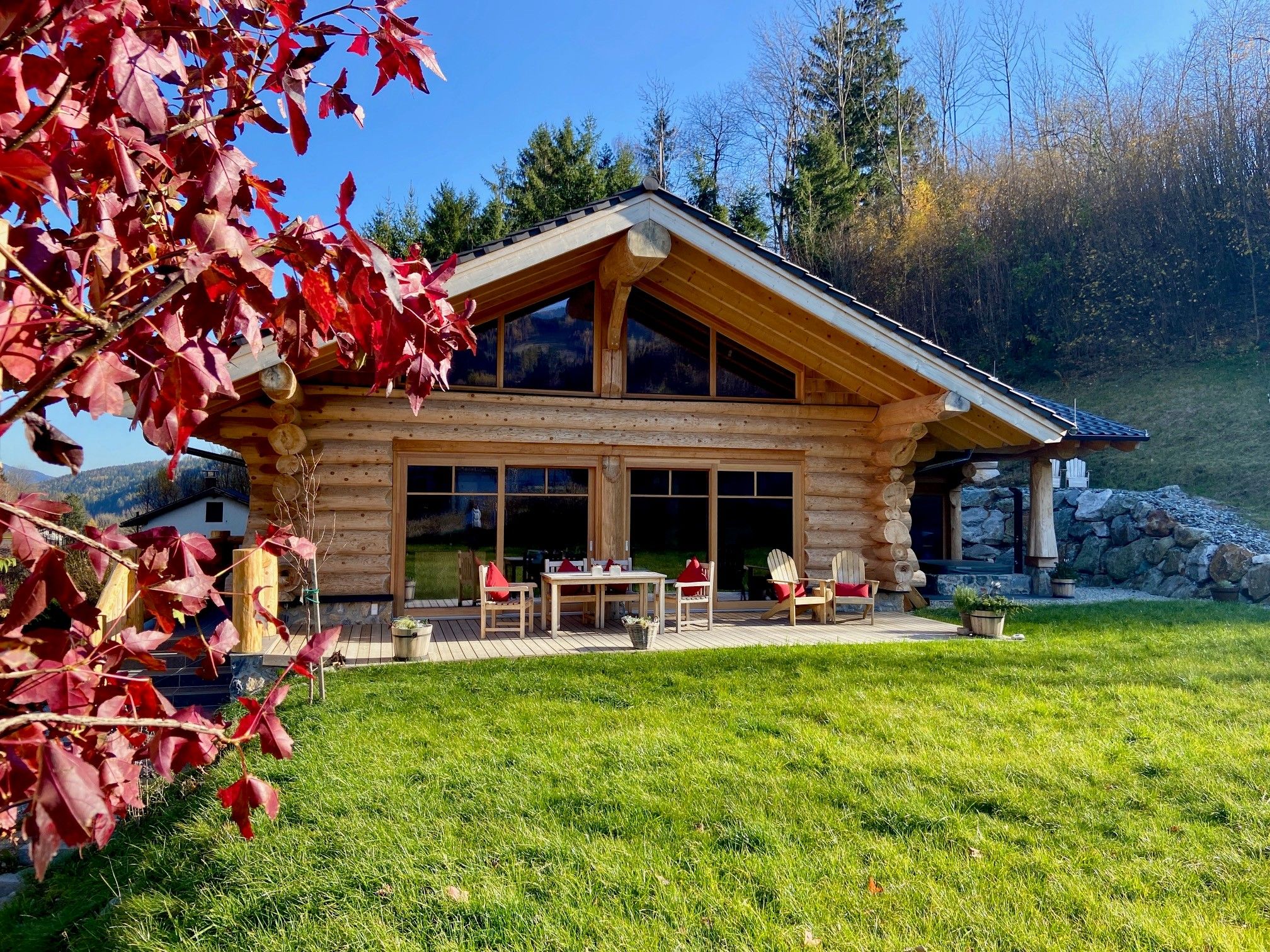 Ein Blockhaus mit großer Terrasse, umgeben von grüner Wiese und herbstlichen Bäumen.
