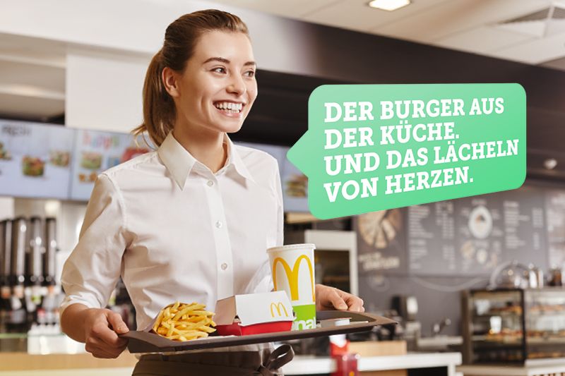 Eine lächelnde Mitarbeiterin in einem Fast-Food-Restaurant serviert ein Tablett mit Pommes, Getränk und Burger. Text: 'Der Burger aus der Küche. Und das Lächeln von Herzen.'