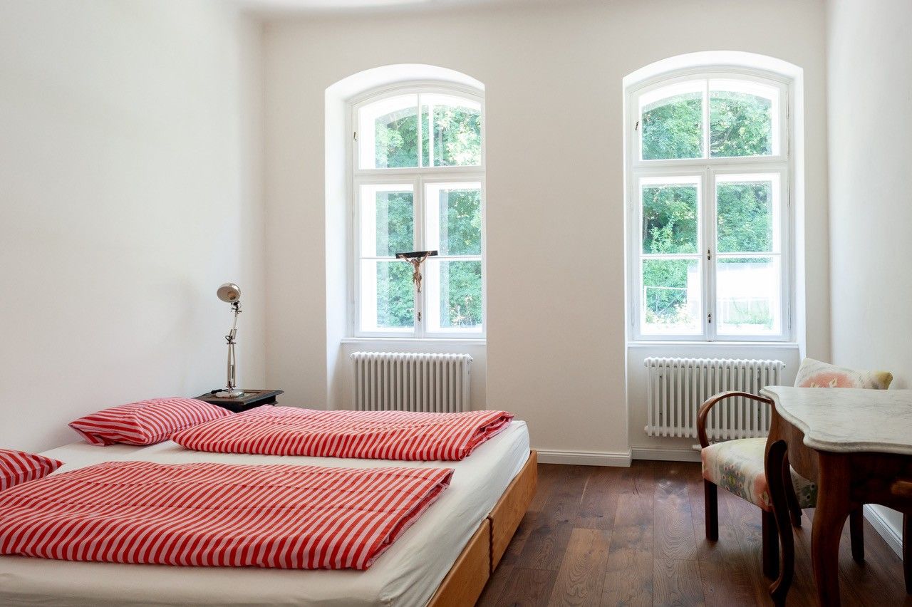 Ein helles Schlafzimmer mit einem Doppelbett, roten gestreiften Bettdecken, zwei großen Fenstern und einem antiken Stuhl mit Tisch.