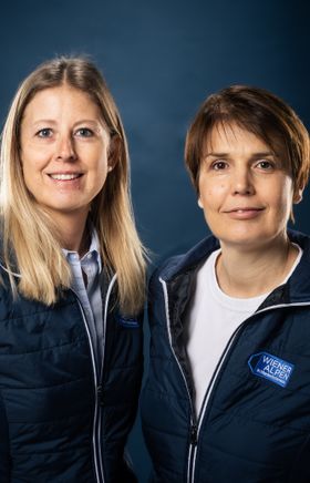Portraitfoto von zwei Wiener Alpen Mitarbeiterinnen, die eine Jacke mit Wiener Alpen Logo tragen und vor einem dunklem Hintergrund stehen.