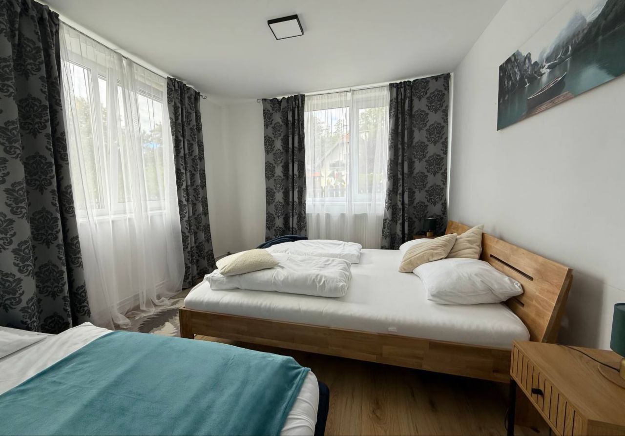 Helles Schlafzimmer mit zwei Betten, Holzboden und großen Fenstern mit Vorhängen.