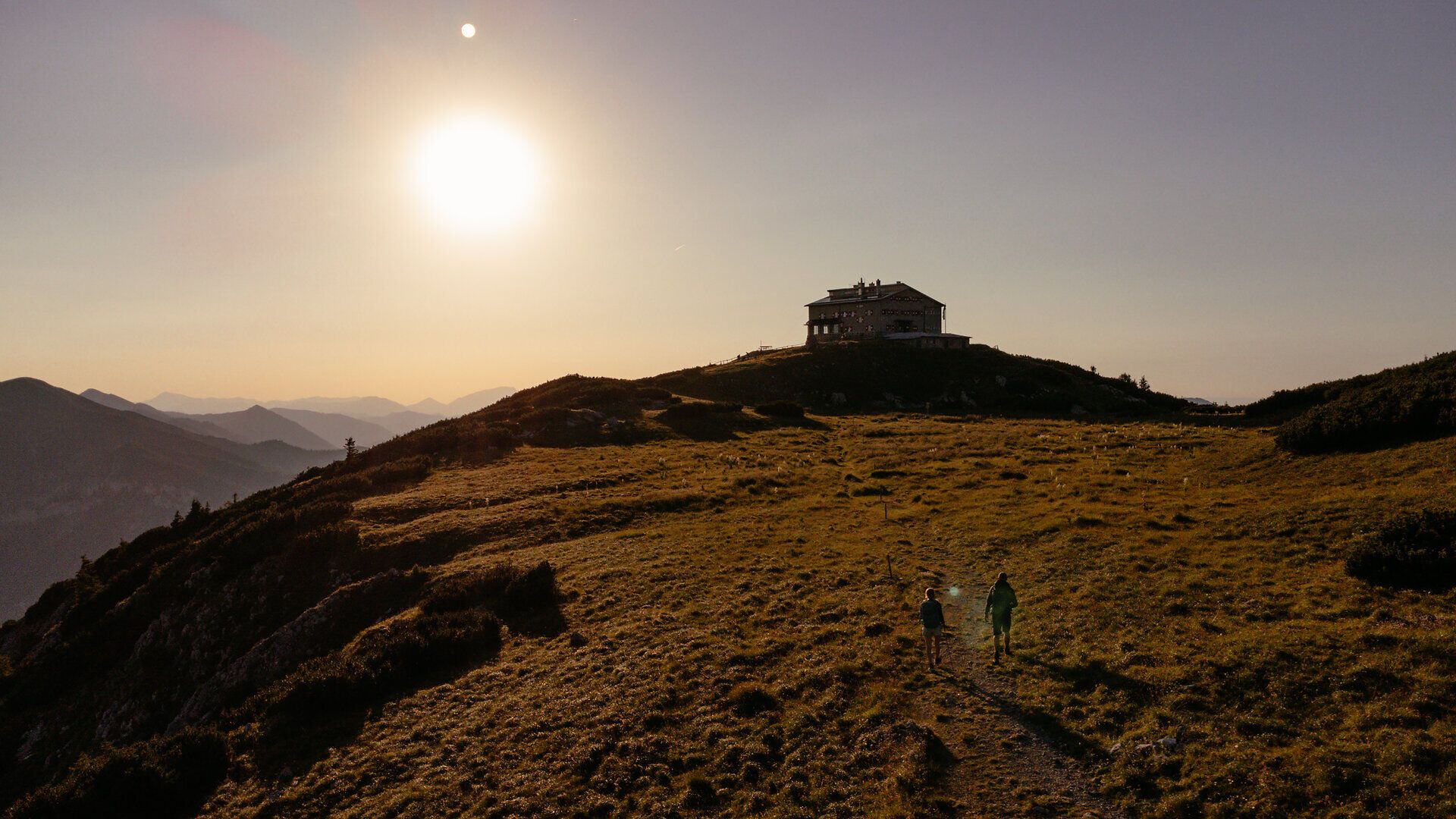 Die sanften Hügel und die goldene Abendsonne schaffen eine magische Atmosphäre, während Wanderer den Pfad entlang der Wiesen erkunden. Die beeindruckende Aussicht auf die umliegenden Berge lädt dazu ein, innezuhalten und die Schönheit der Natur zu genießen.