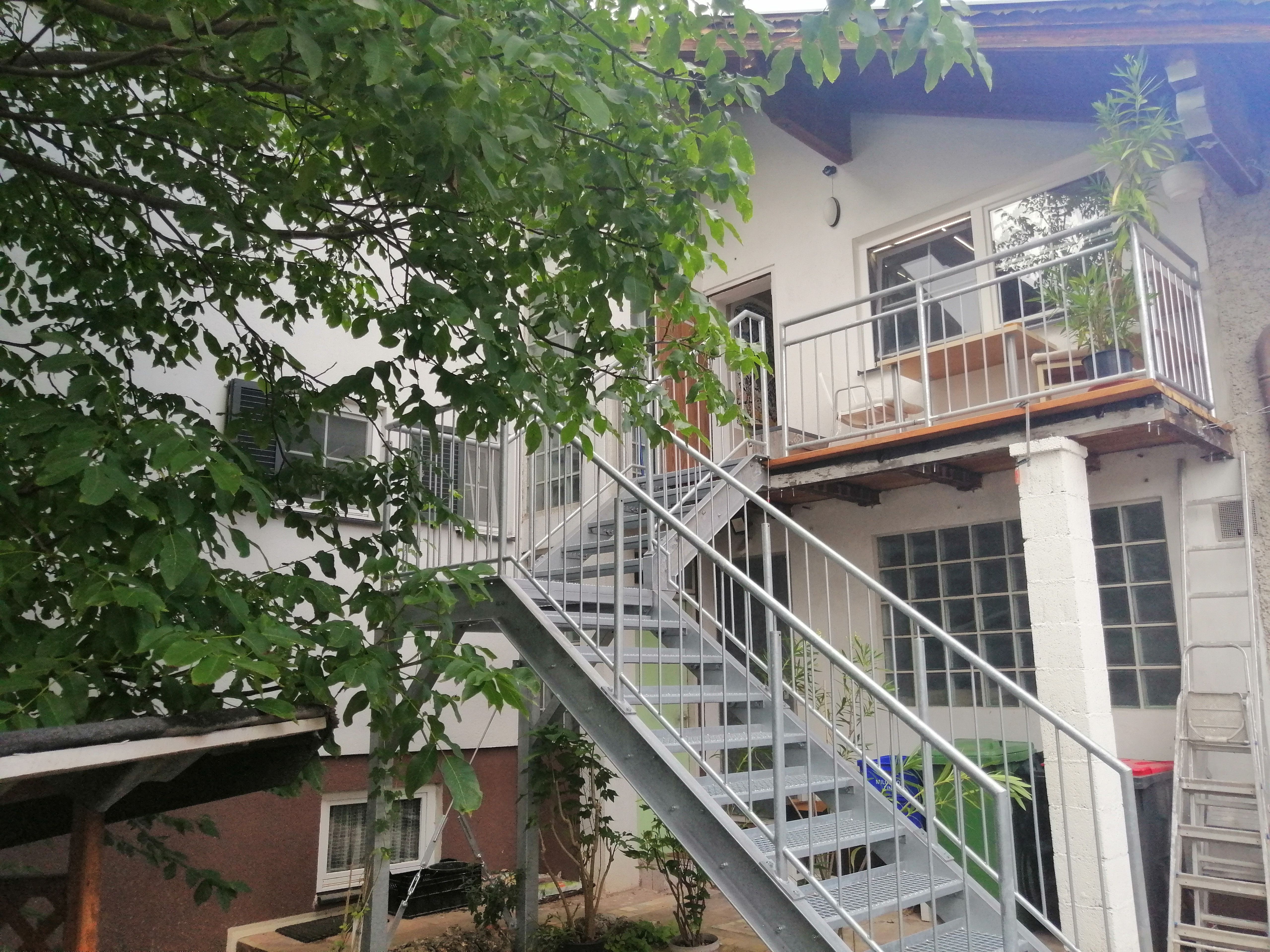Außenansicht eines Gebäudes mit Metalltreppe und Balkon, umgeben von einem Nussbaum.