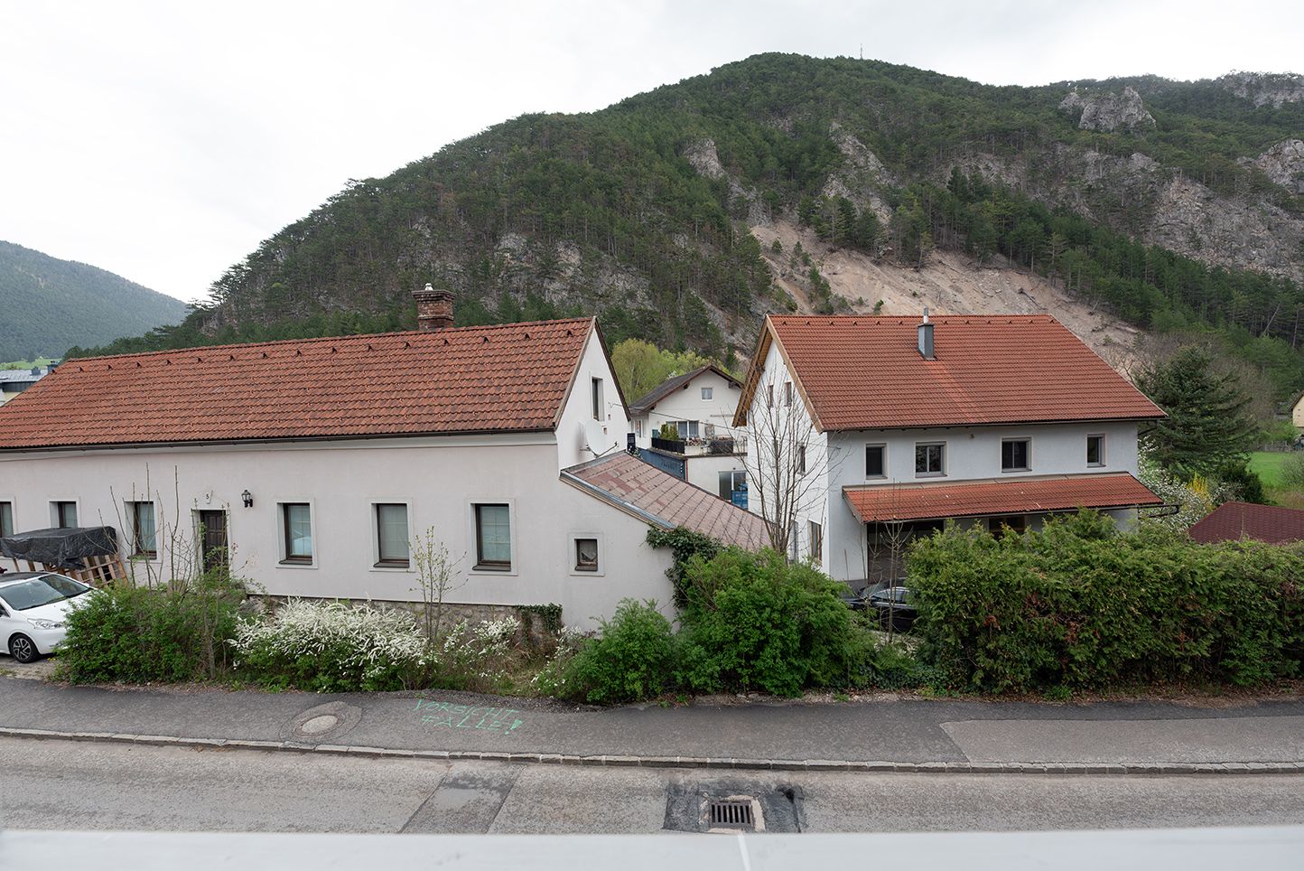 Zwei Häuser mit roten Dächern vor einem bewaldeten Hügel.