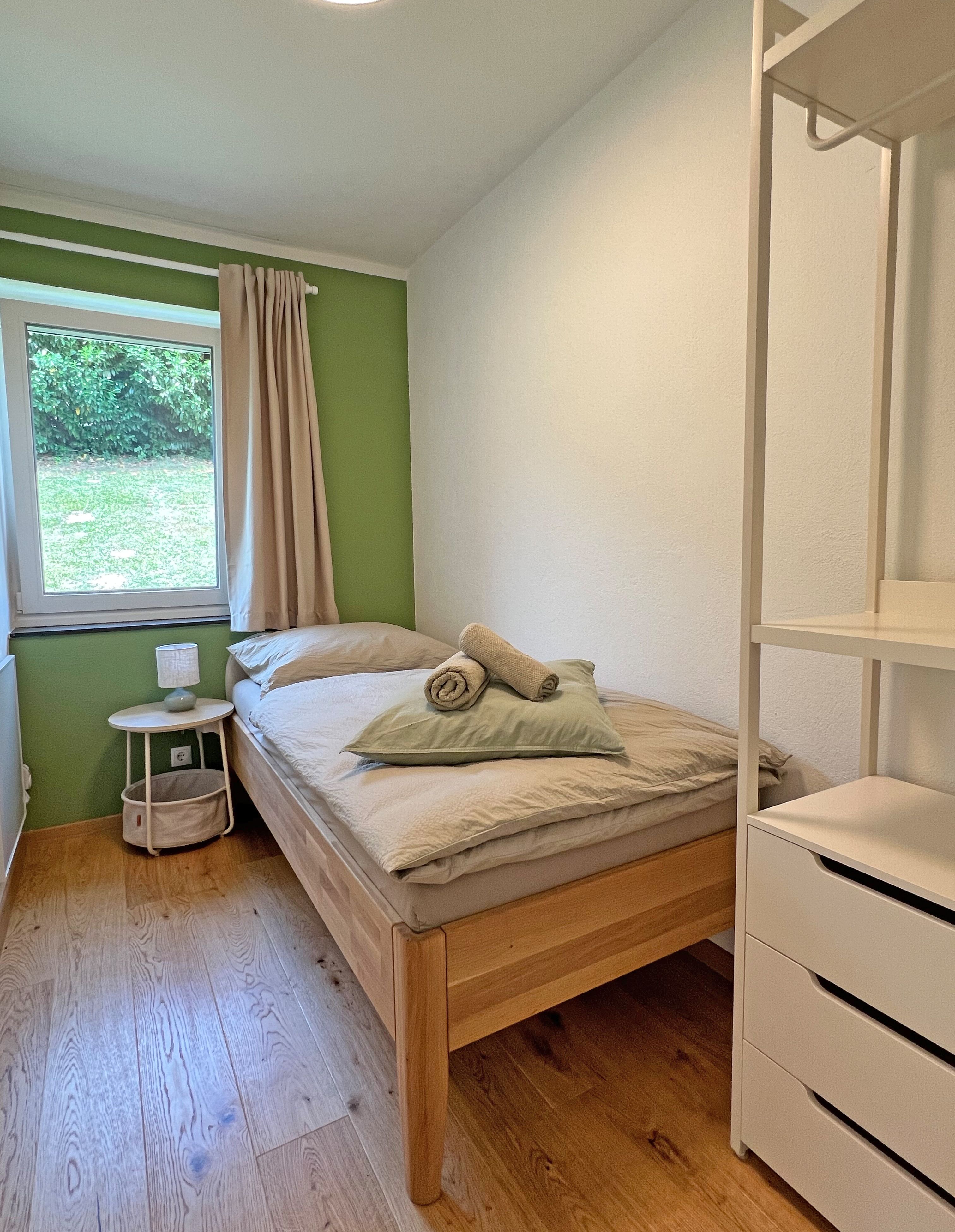 A small bedroom with green-and-white walls, a single bed, a nightstand, a window with a curtain, and a white shelf.