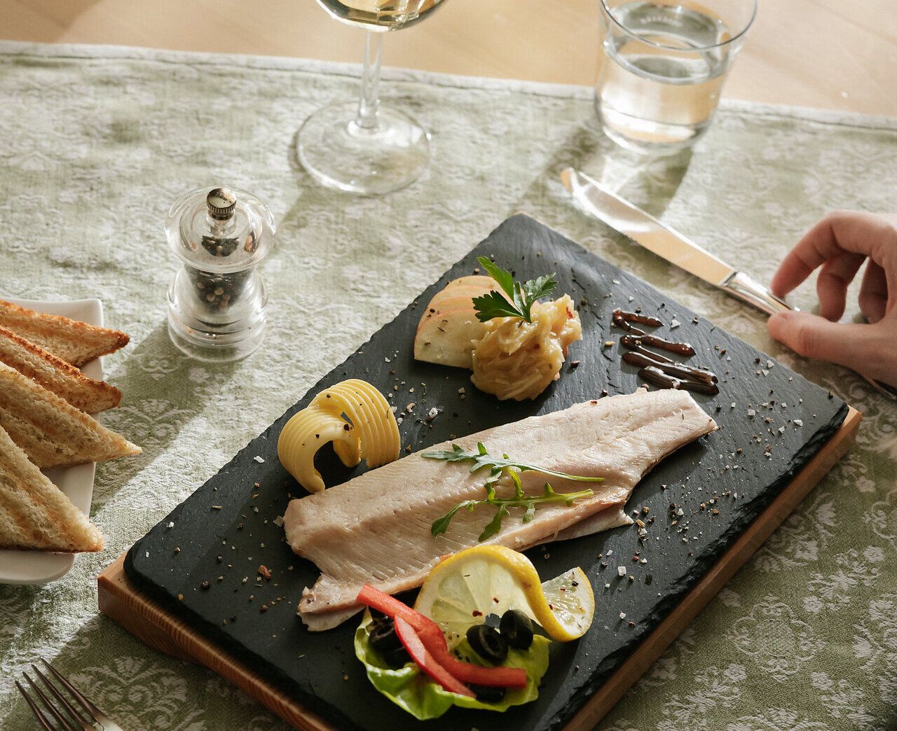 Angerichteter Teller mit geräuchertem Fisch mit Garnierung, dahinter steht ein Glas Wein und auf einem Teller liegen getoastete Brotscheiben.