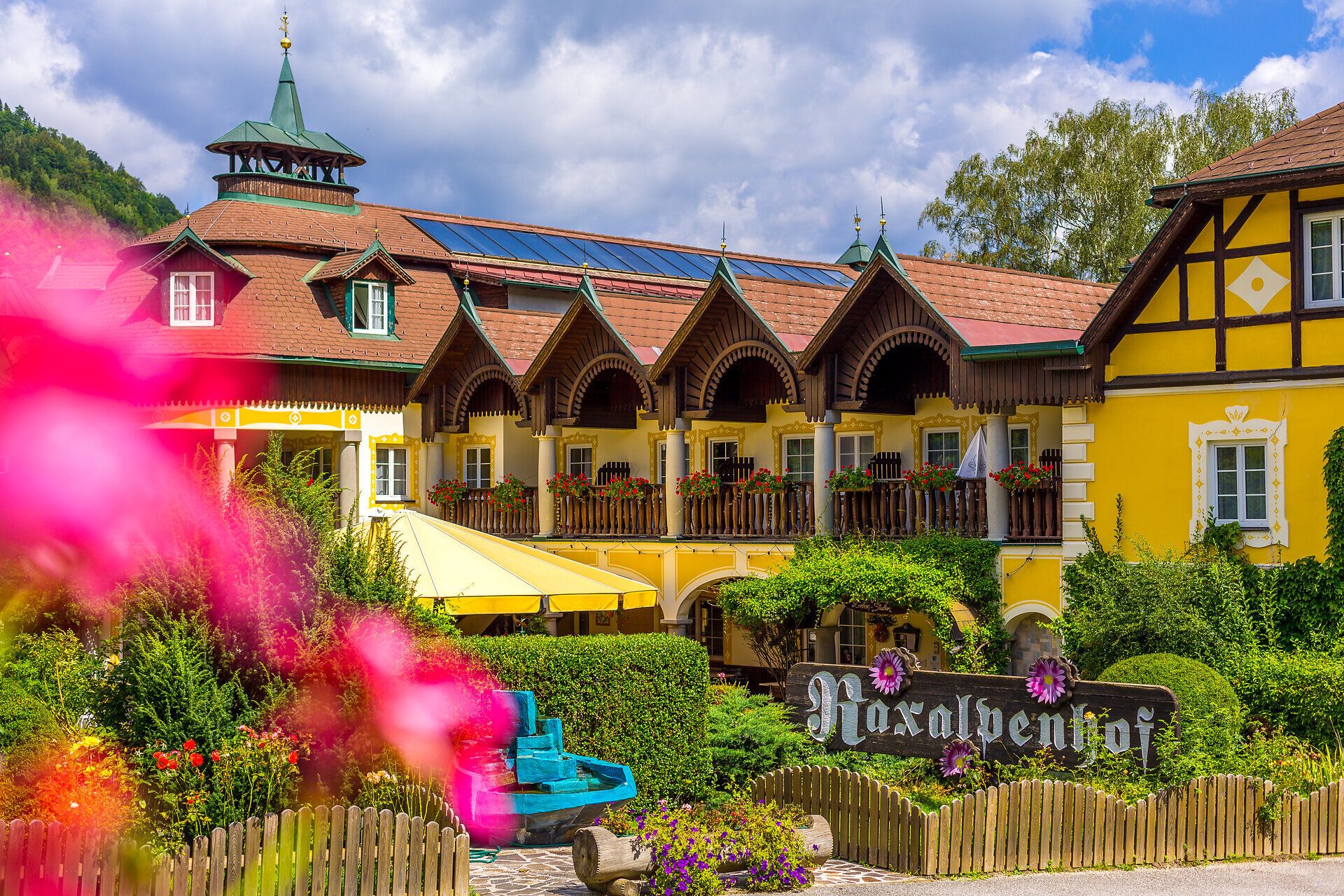 Raxalpenhof liegt direkt an der Ortsstraße, vor dem Haus steht ein blauer Brunnen, Blumen und Sträucher säumen die Balkone und en gesamten Vorderbereich des Hauses