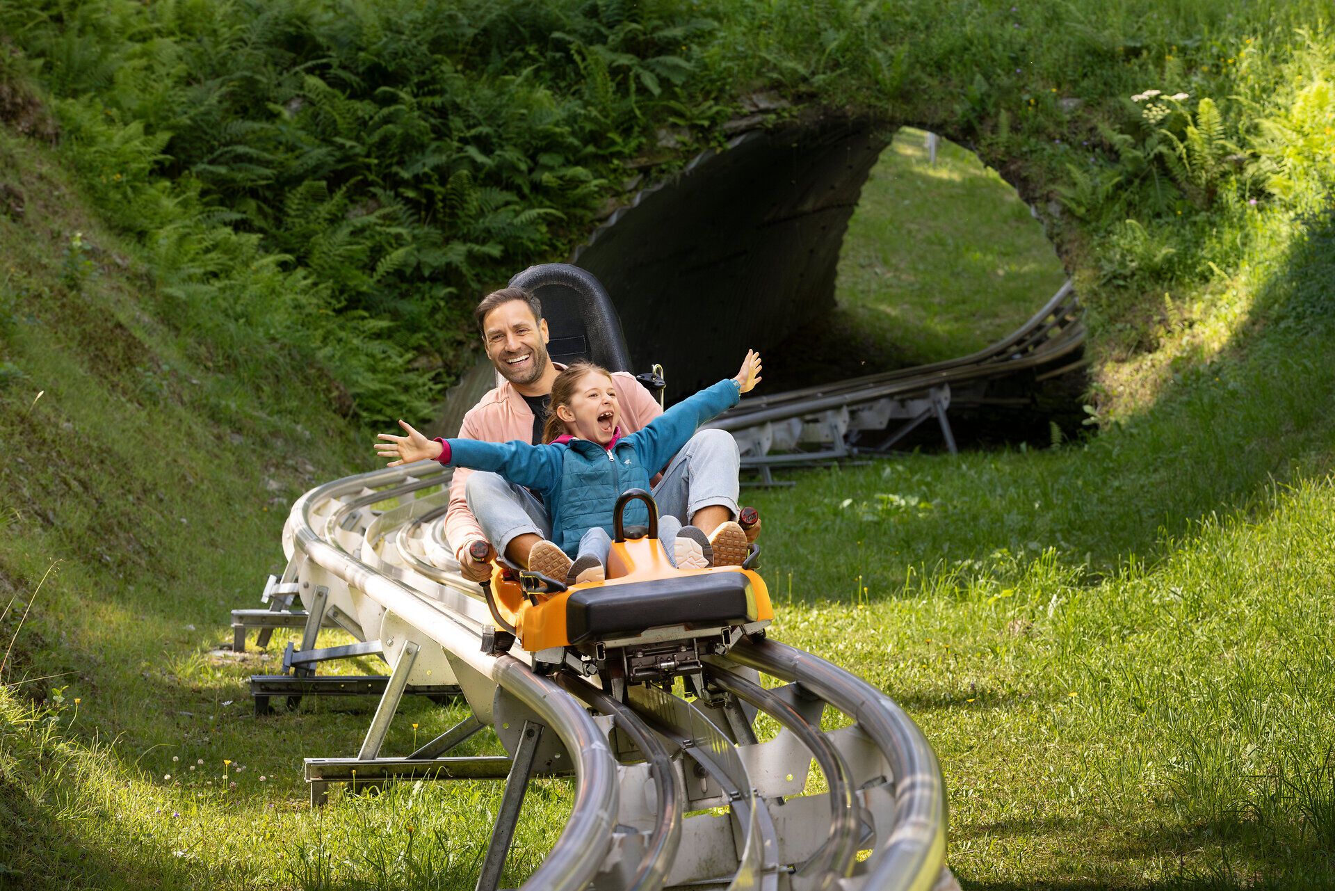 Ein fröhlicher Ausblick auf ein aufregendes Sommerabenteuer, während ein Vater mit seinem Kind auf der Sommerrodelbahn den Hang hinunter saust. Die strahlenden Gesichter und die umgebende grüne Landschaft schaffen eine unvergessliche Atmosphäre voller Freude und Freiheit.