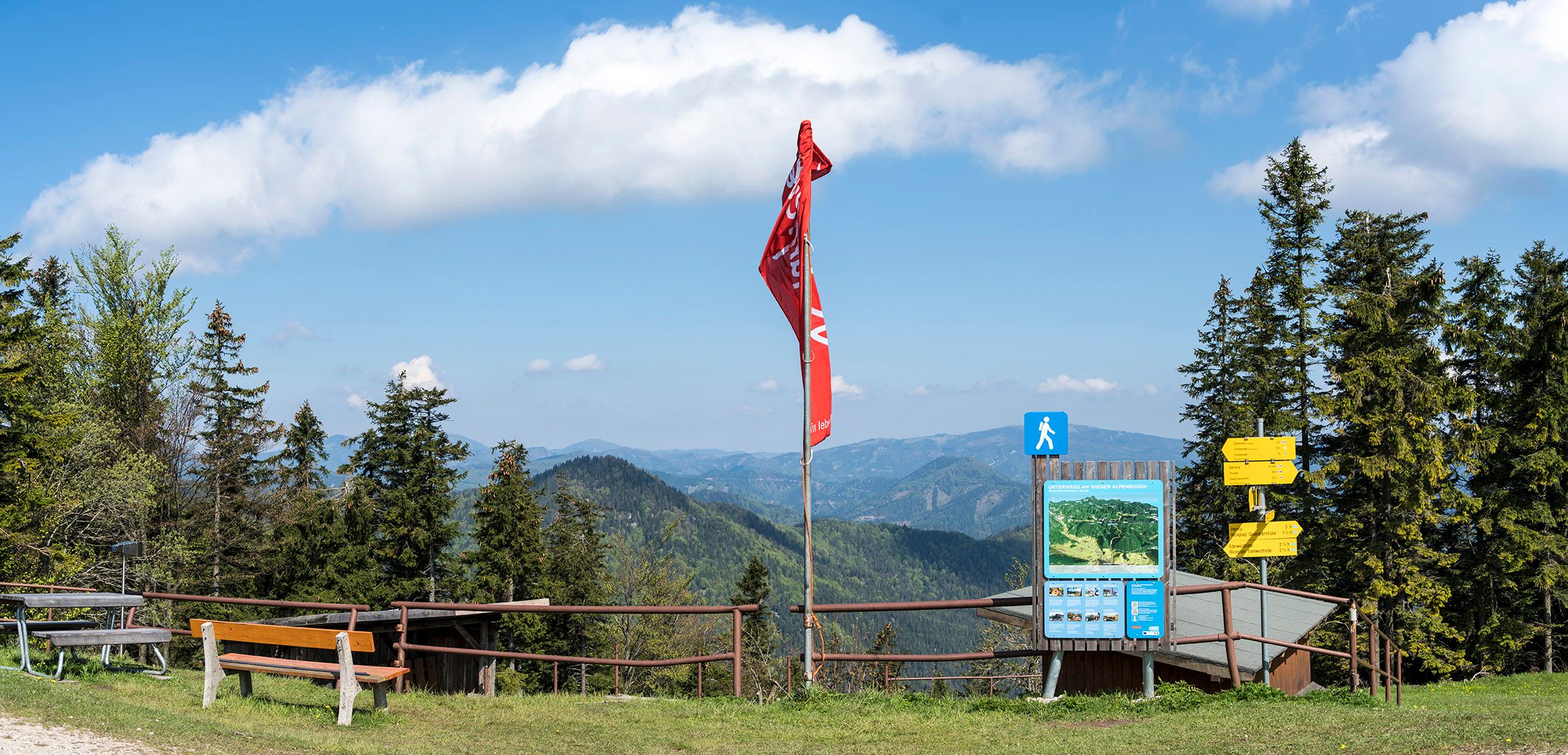 Aussichtspunkt mit Bank, roter Fahne und Wegweisern vor Berglandschaft.