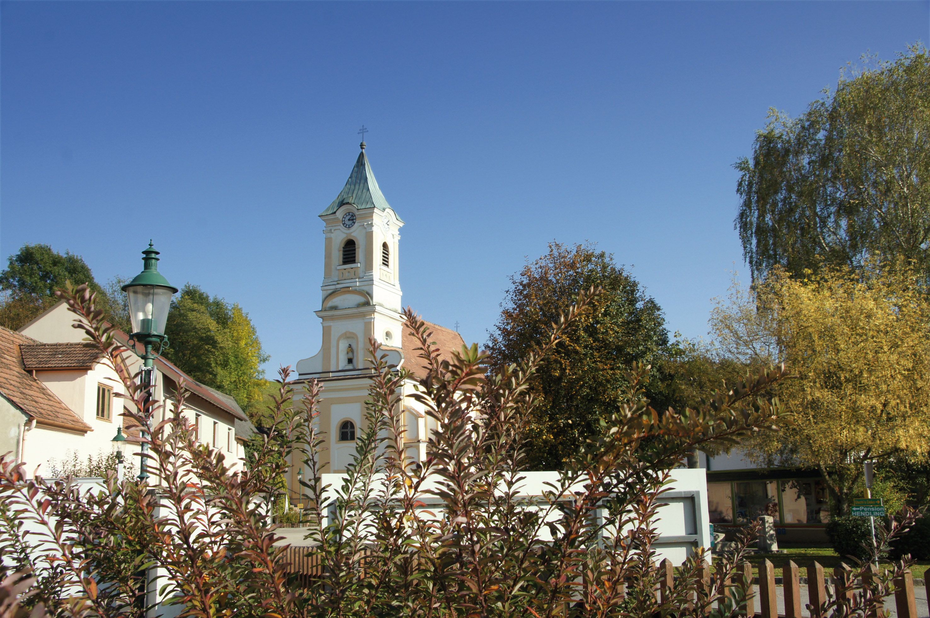 Pfarrkirche Walpersbach mit Turm und umliegenden Gebäuden, umgeben von Bäumen und Sträuchern unter blauem Himmel.