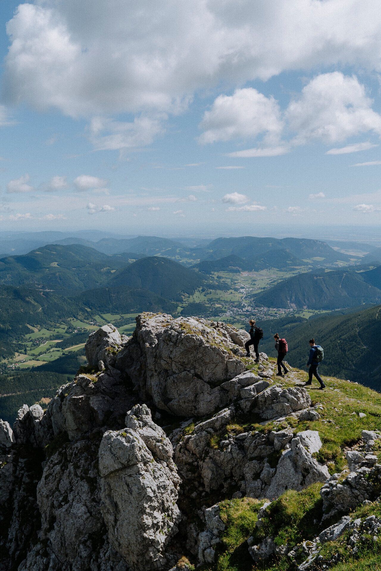 Drei Wandernde erklimmen einen felsigen Gipfel, von dem aus ein beeindruckender Blick über ein weitläufiges Bergpanorama und grüne Täler unter einem blauen Himmel mit weißen Wolken geboten wird, was ein Gefühl von Freiheit und Abenteuer vermittelt.