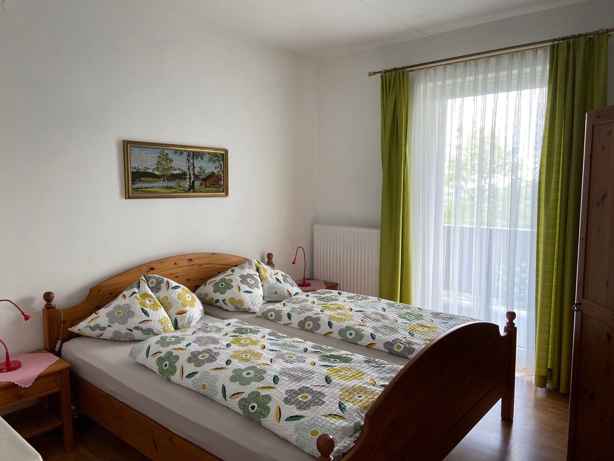 Ein gemütliches Doppelzimmer mit Holzmöbeln, einem großen Fenster mit grünen Vorhängen und einem Landschaftsbild an der Wand.