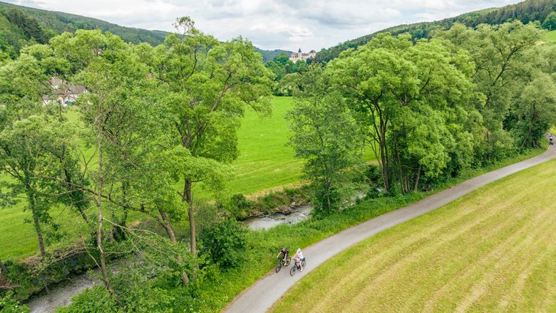 Zwei Radfahrer auf einem Weg entlang eines Flusses, umgeben von grünen Bäumen und Wiesen.