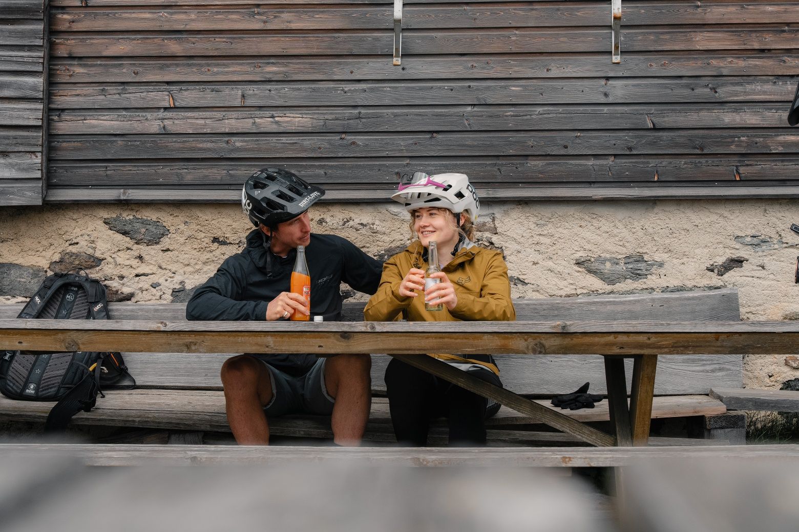 Zwei Mountainbiker sitzen auf einer Holzbank vor einer Hütte und trinken Erfrischungsgetränke.