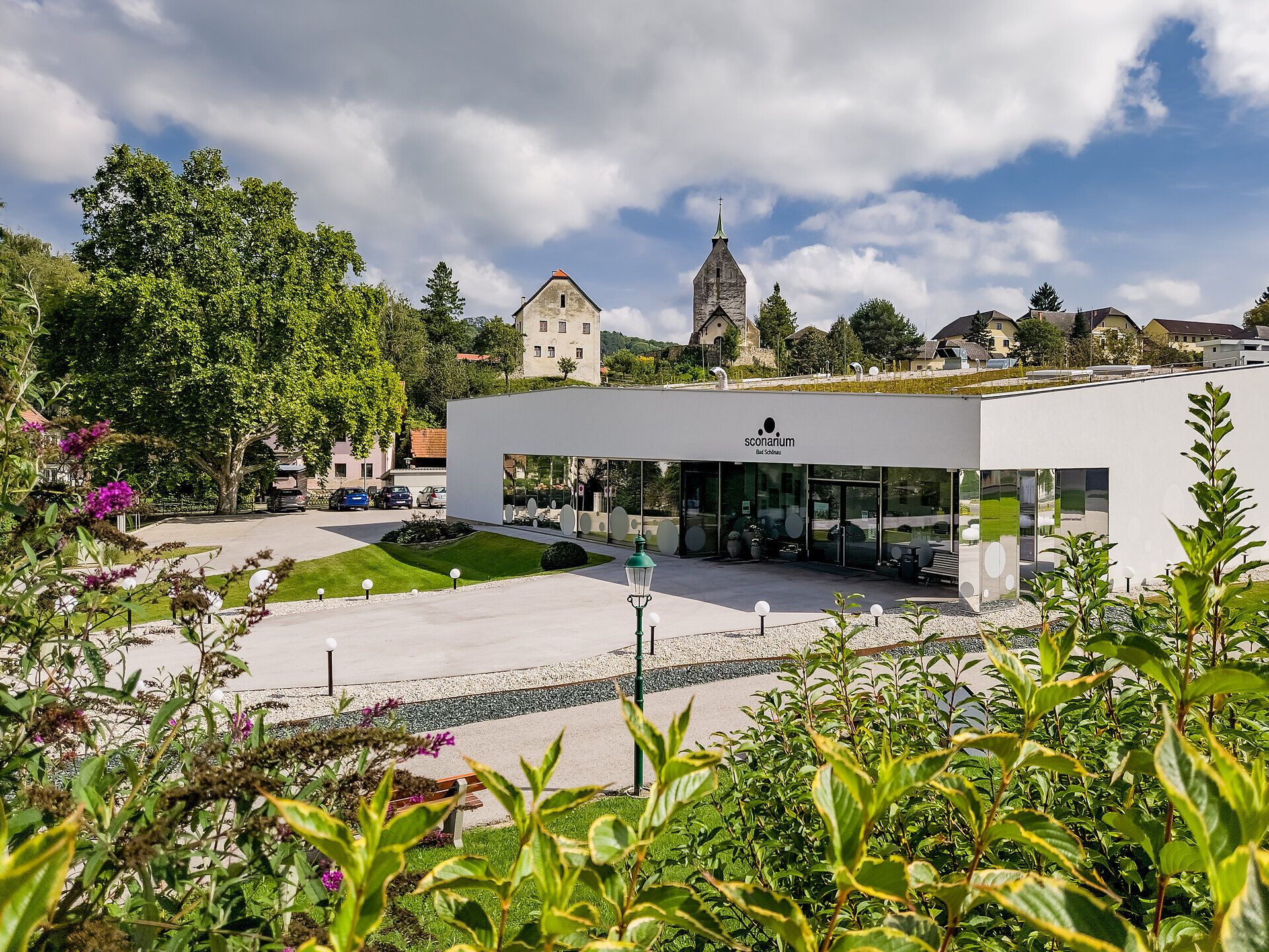 Umgeben von sanften Hügeln und üppigem Grün strahlt das Sconarium Bad Schönau eine einladende Ruhe aus. Die moderne Architektur harmoniert perfekt mit der malerischen Landschaft der Buckligen Welt, während die Wolken über den Himmel ziehen und die Atmosphäre mit einem Hauch von Abenteuer erfüllen.