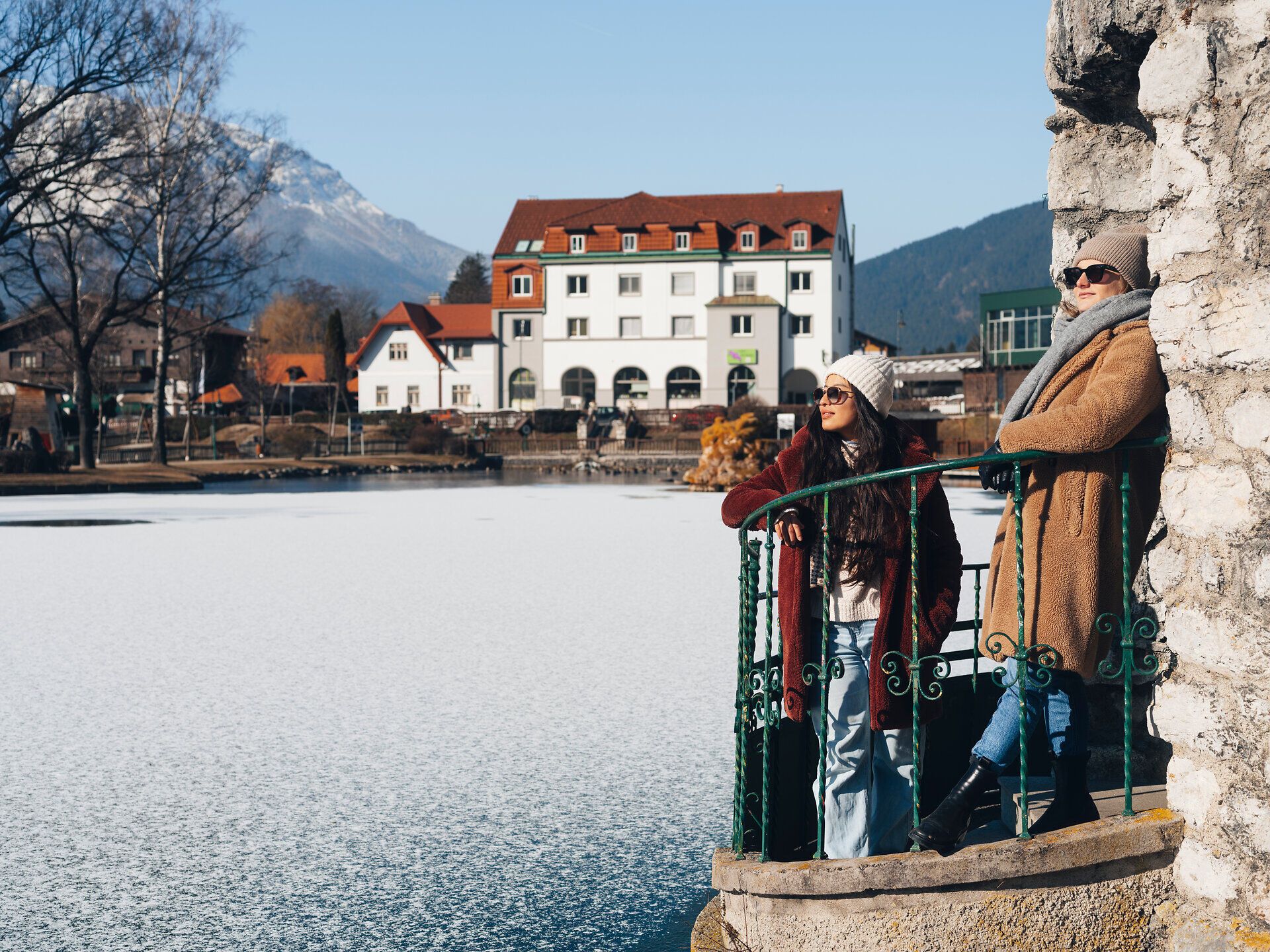 An einem klaren Wintertag genießen zwei Frauen die friedliche Atmosphäre am Großen Teich. Die schneebedeckten Berge im Hintergrund und die glitzernde Eisfläche schaffen eine malerische Kulisse, die zum Verweilen einlädt.