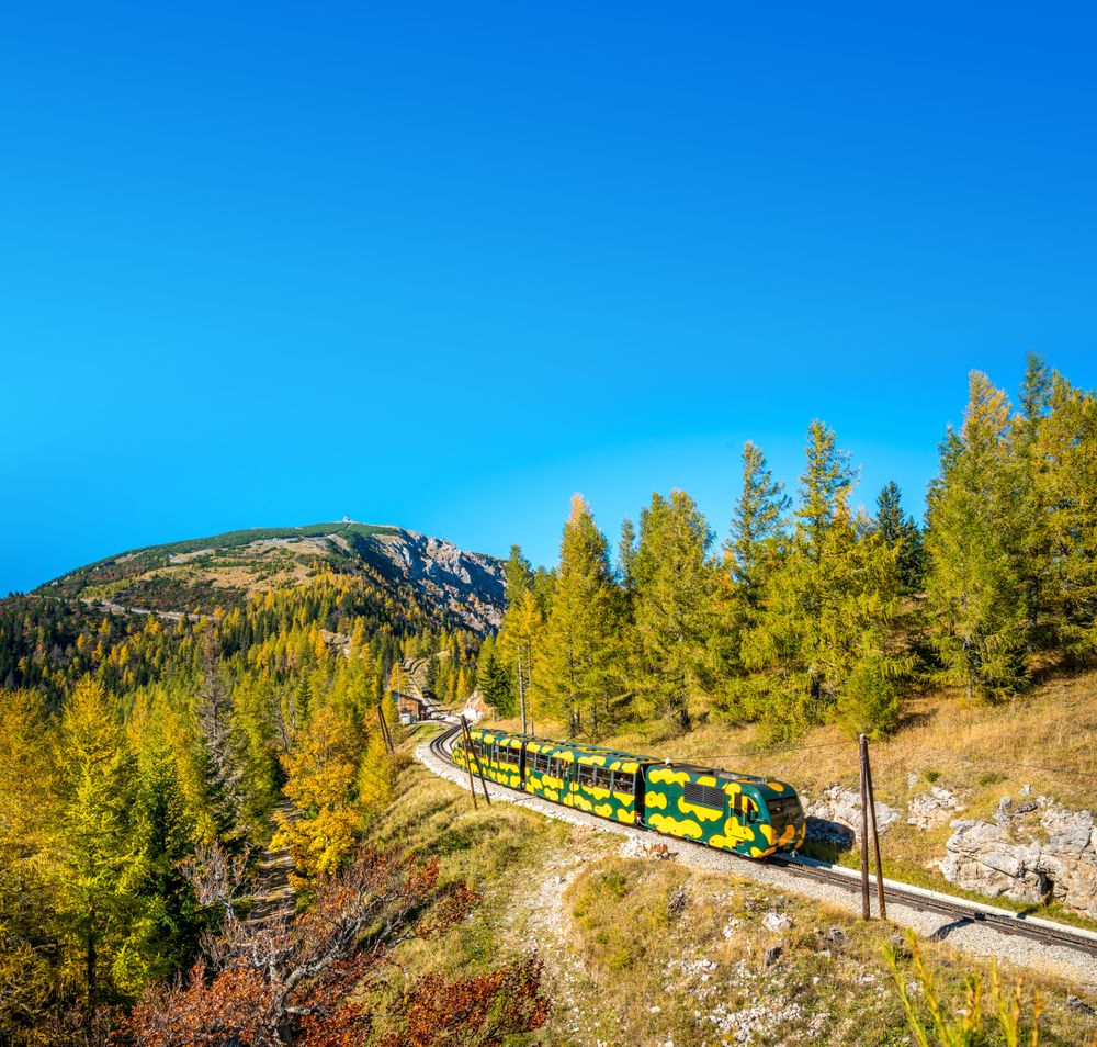A train in salamander design travels through an autumnal mountain landscape with a blue sky.
