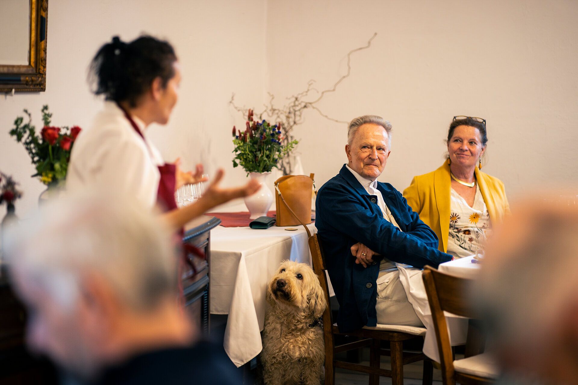 Eine Person steht in einer Gaststube und spricht mit zwei am Tisch sitzenden Personen. Ein Hund sitzt daneben und schaut die stehende Person an.