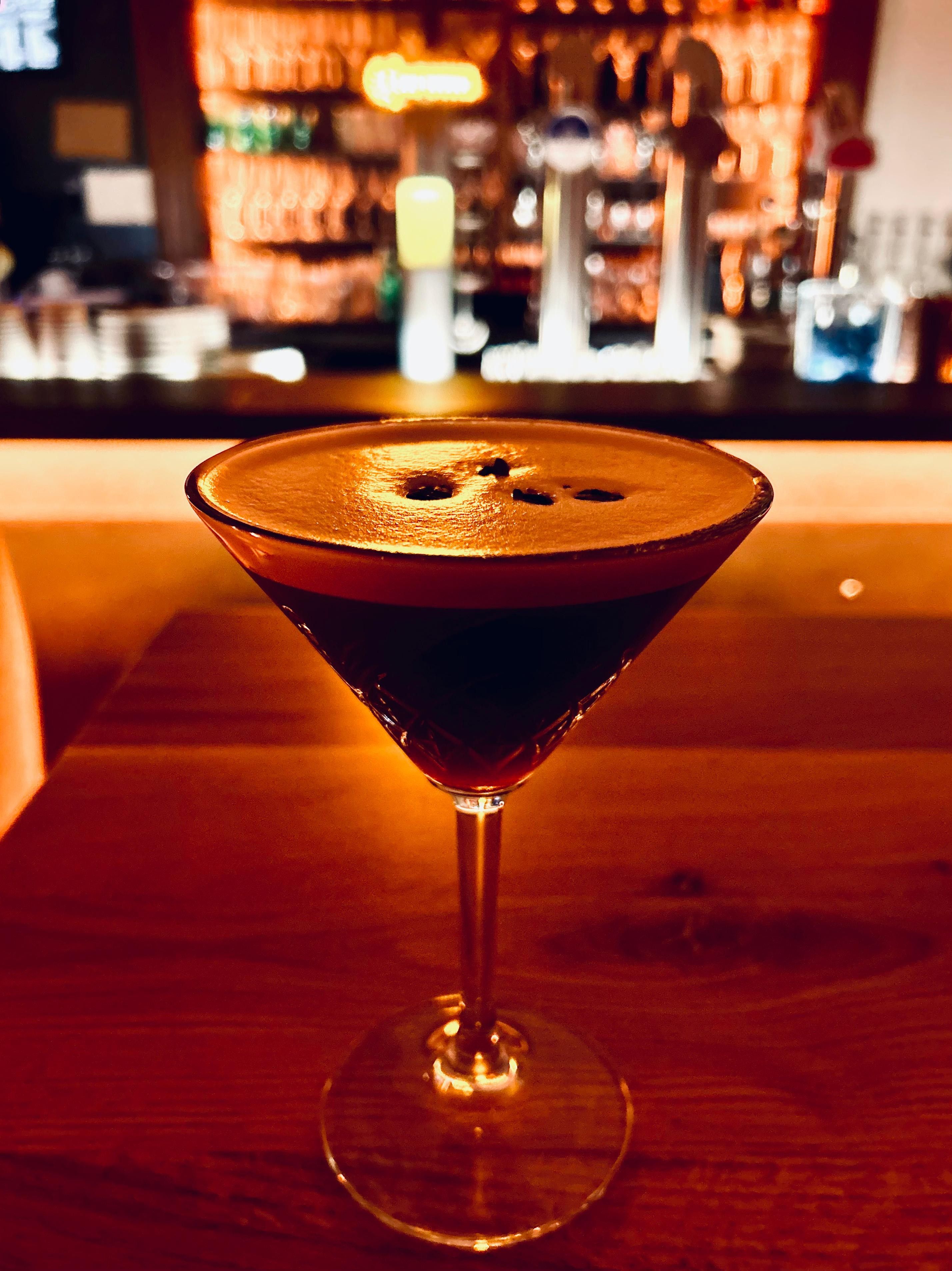 Ein Cocktailglas mit dunklem Getränk auf einem Holztisch, im Hintergrund eine unscharfe Bar.