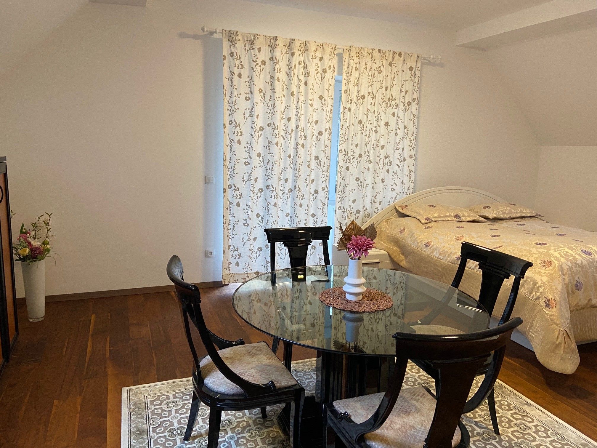 Ein Wohn-Schlafzimmer mit einem runden Glastisch, vier Stühlen, einem Bett und Vorhängen mit Blumenmuster.