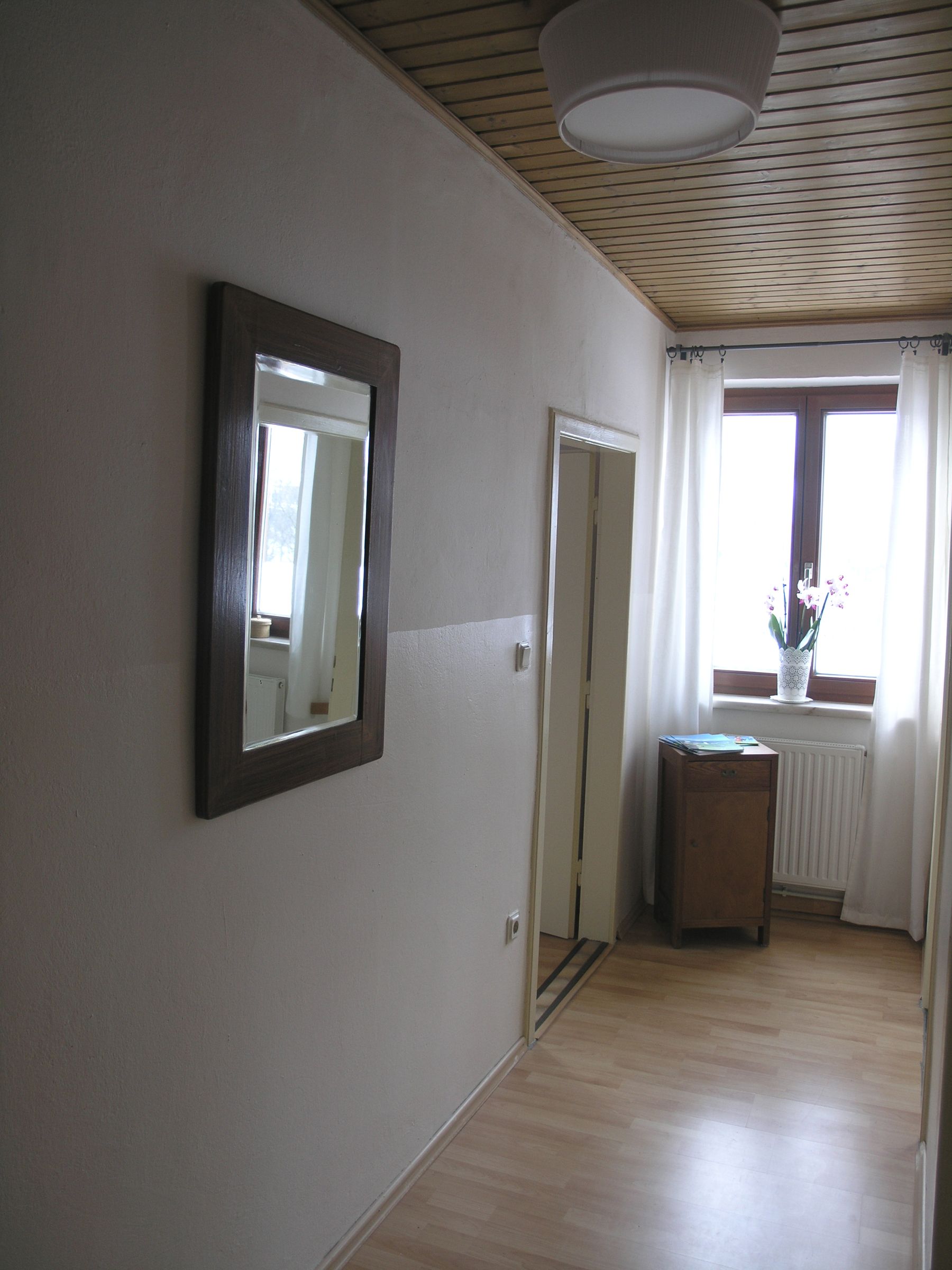 Ein schmaler Flur mit Holzdecke, Spiegel an der Wand, Fenster mit Vorhängen und kleinem Tisch mit Pflanze.
