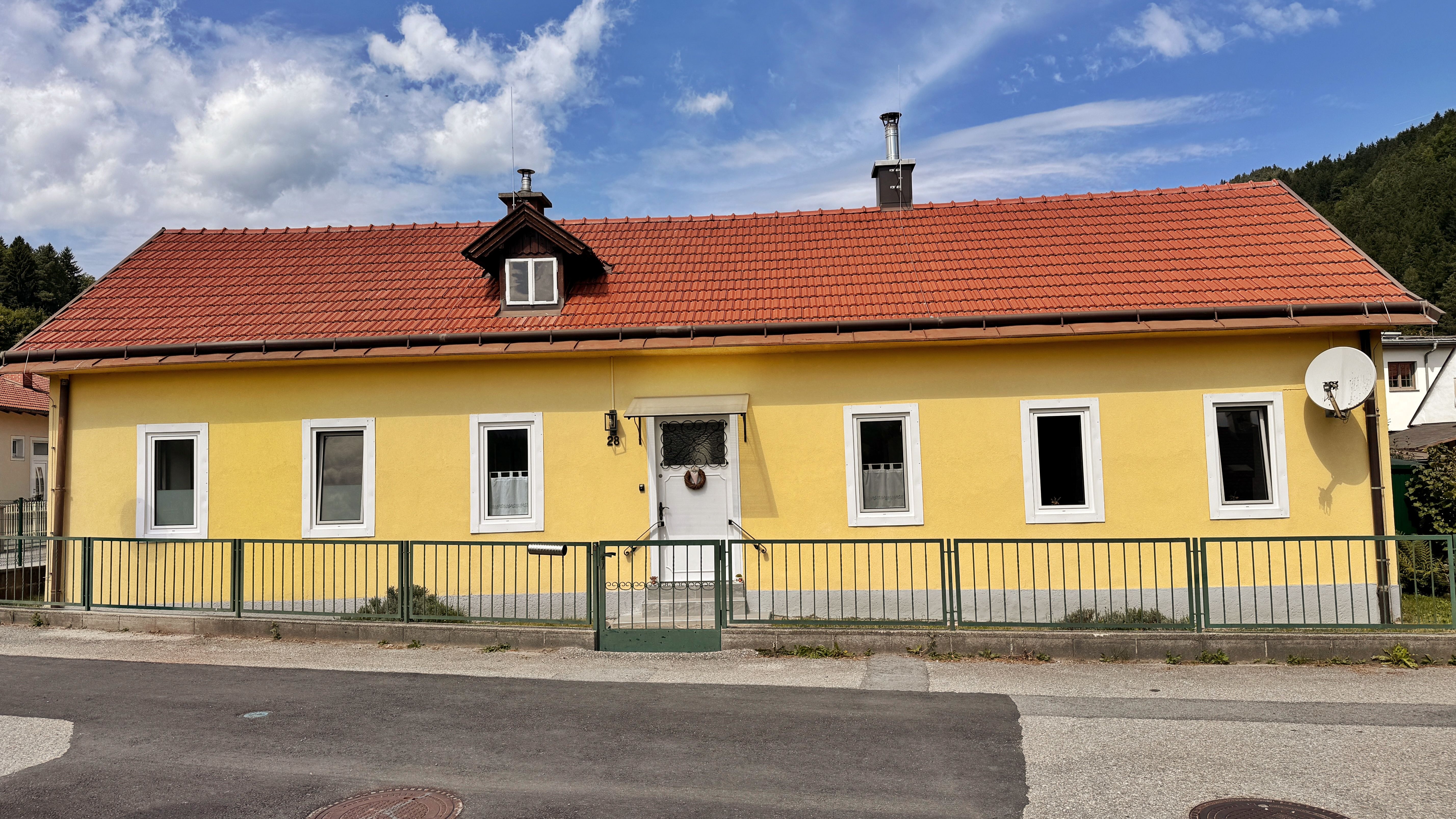 Gelbes Haus mit rotem Dach und mehreren Fenstern, umgeben von einem grünen Zaun.