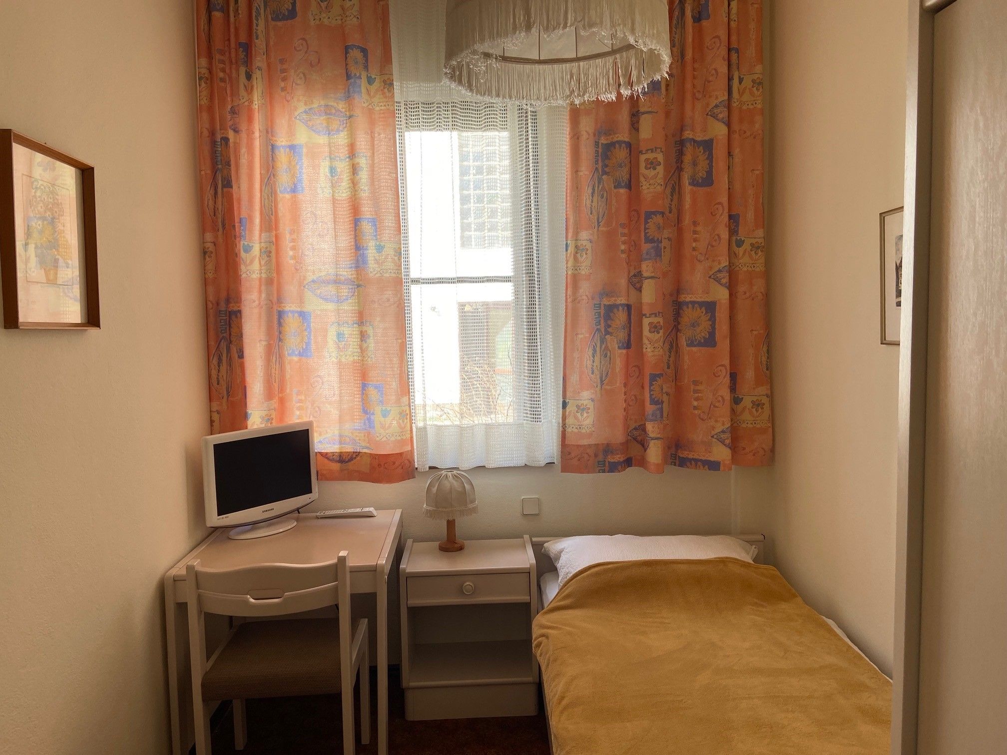 Ein kleines Einzelzimmer mit Bett, Schreibtisch, Stuhl und Fernseher. Bunte Vorhänge und eine Lampe sind ebenfalls zu sehen.