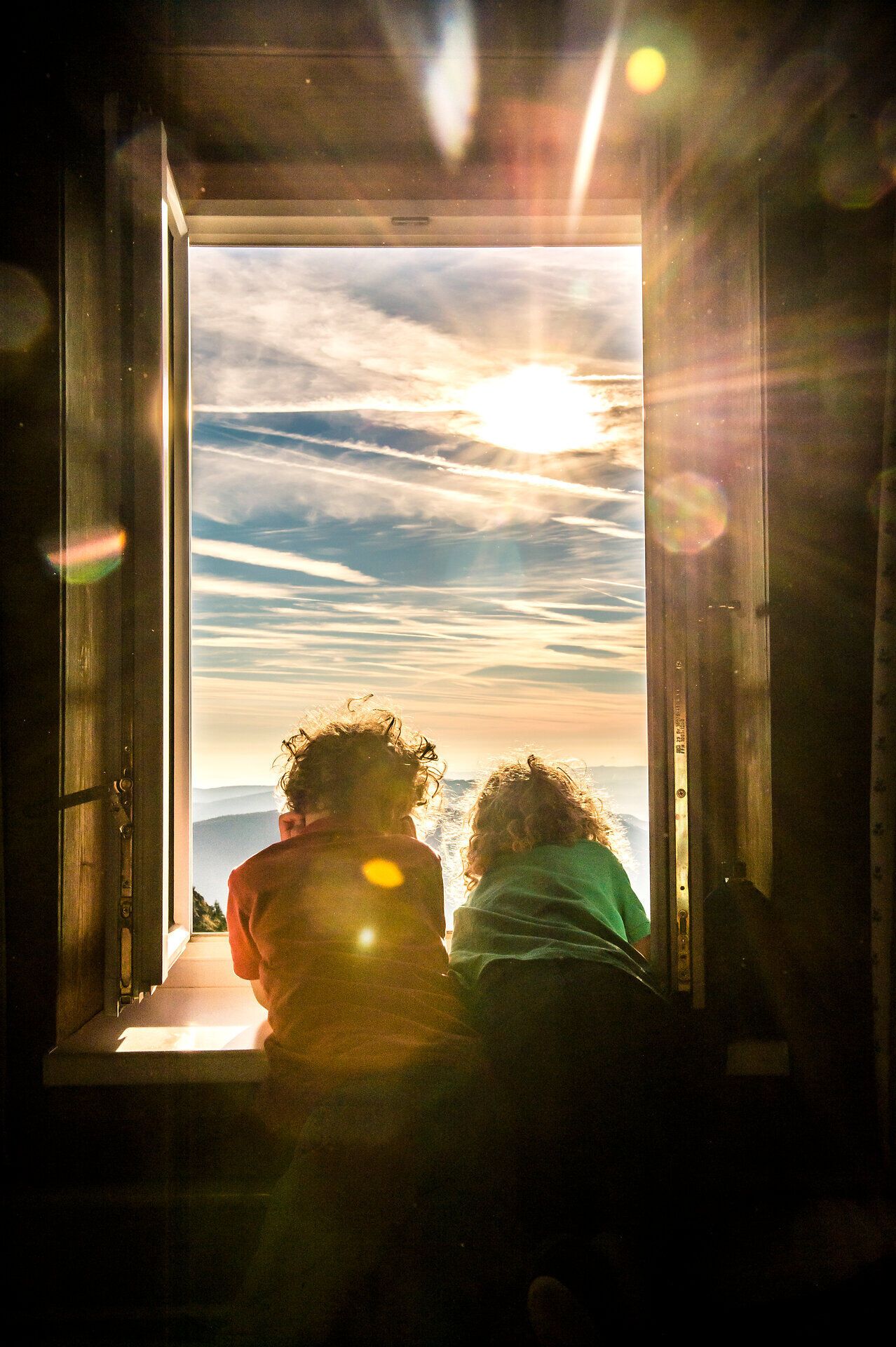 Zwei Kinder sitzen am Fenster und blicken fasziniert in die Ferne, während die Sonne sanft über die Berge aufgeht. Die goldenen Strahlen tauchen die Landschaft in ein warmes Licht und schaffen eine magische Atmosphäre, die den Sommer in den Bergen perfekt einfängt.