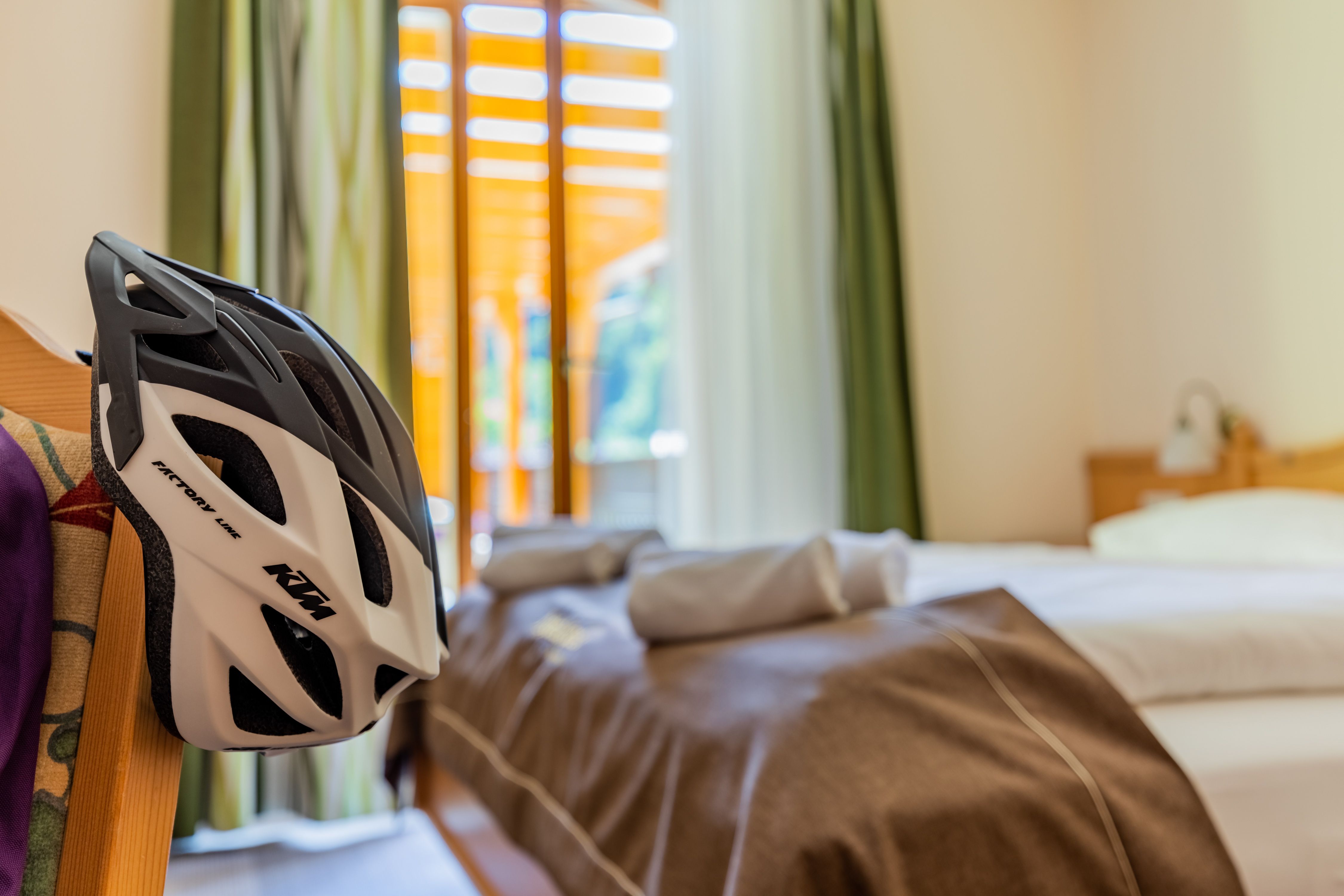 Ein Stuhl auf dem ein Radhelm hängt, im Hintergrund ein Hotelzimmer mit Bett.
