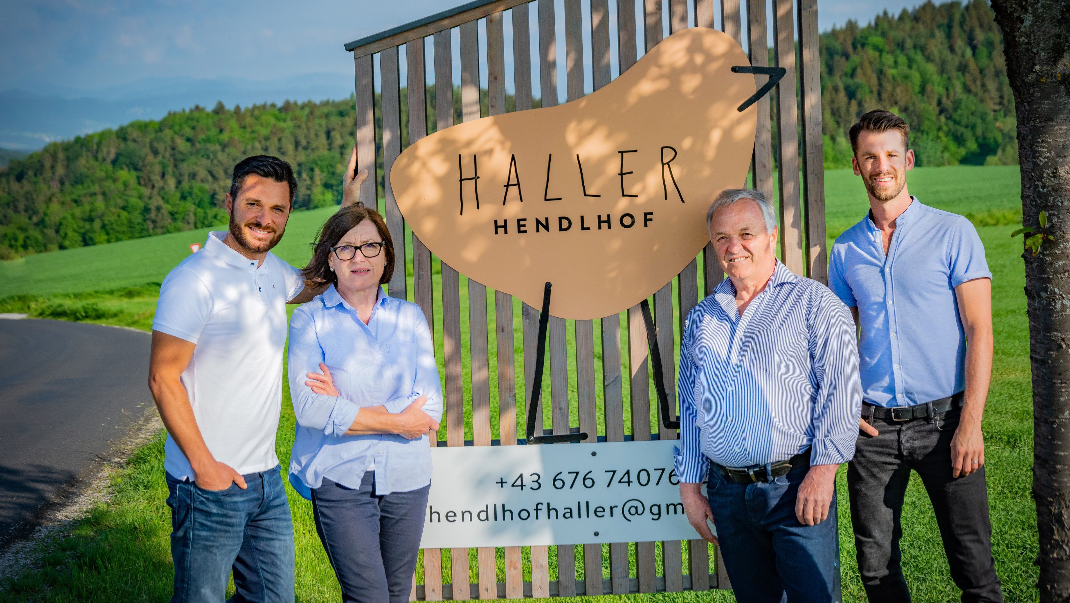 Vier Personen stehen vor einer Holztafel mit der Aufschrift 'Haller Hendlhof' im Freien.