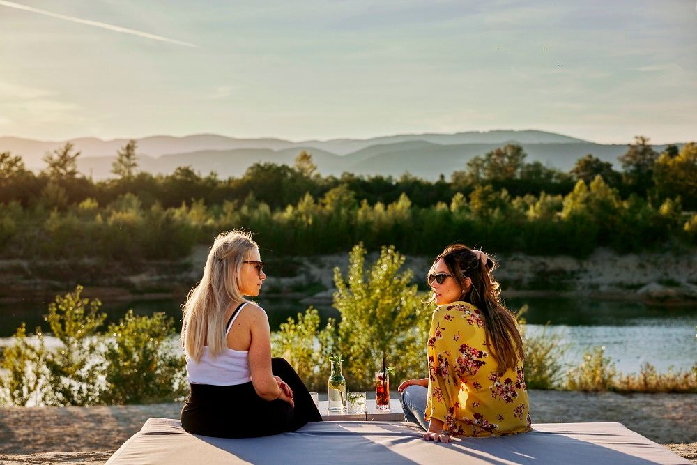 Zwei Frauen sitzen auf einer Decke am See, umgeben von Natur, bei Sonnenuntergang.