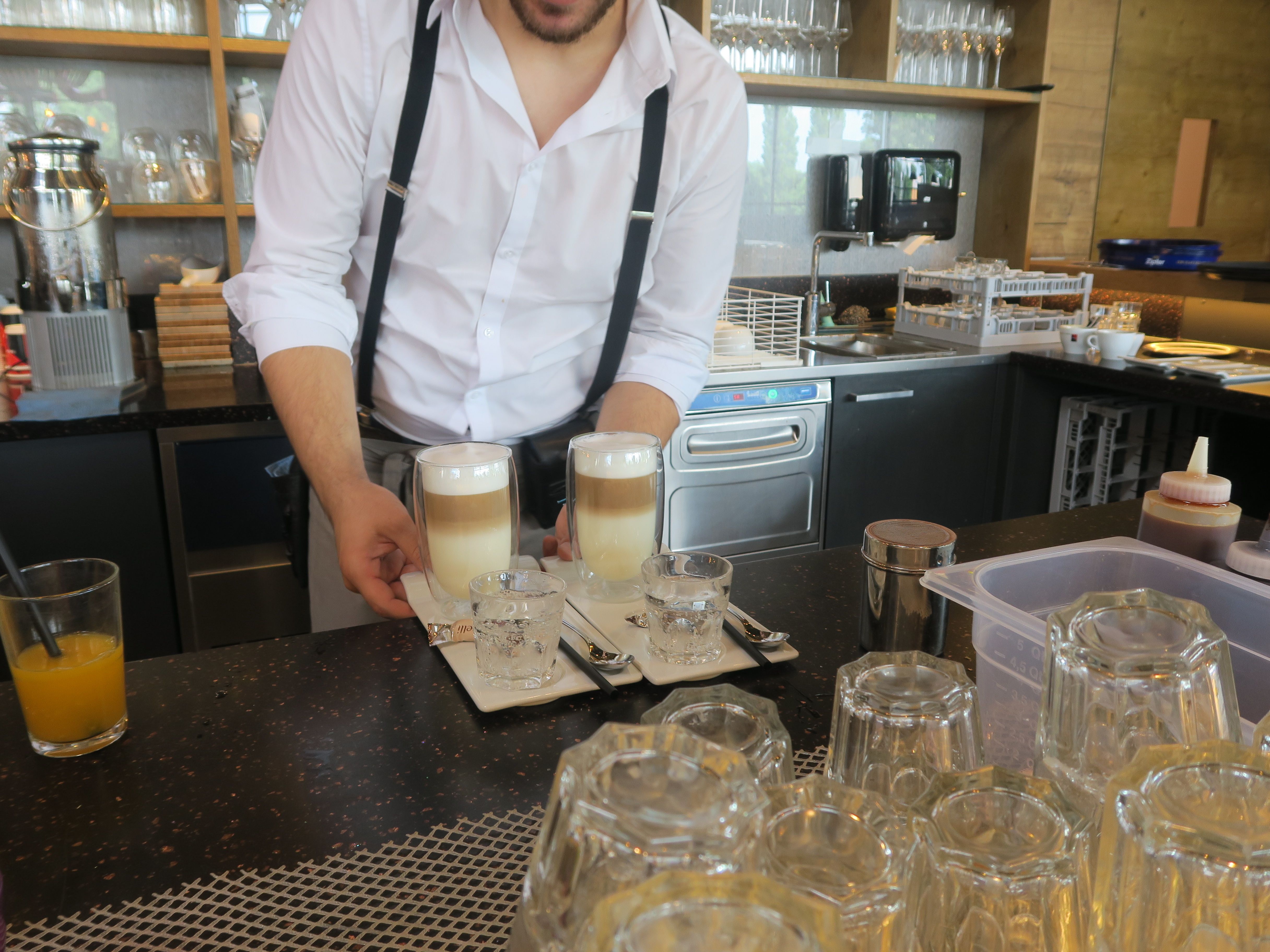 Barista serviert zwei Latte Macchiato mit Wassergläsern auf einem Tablett in einem Café.