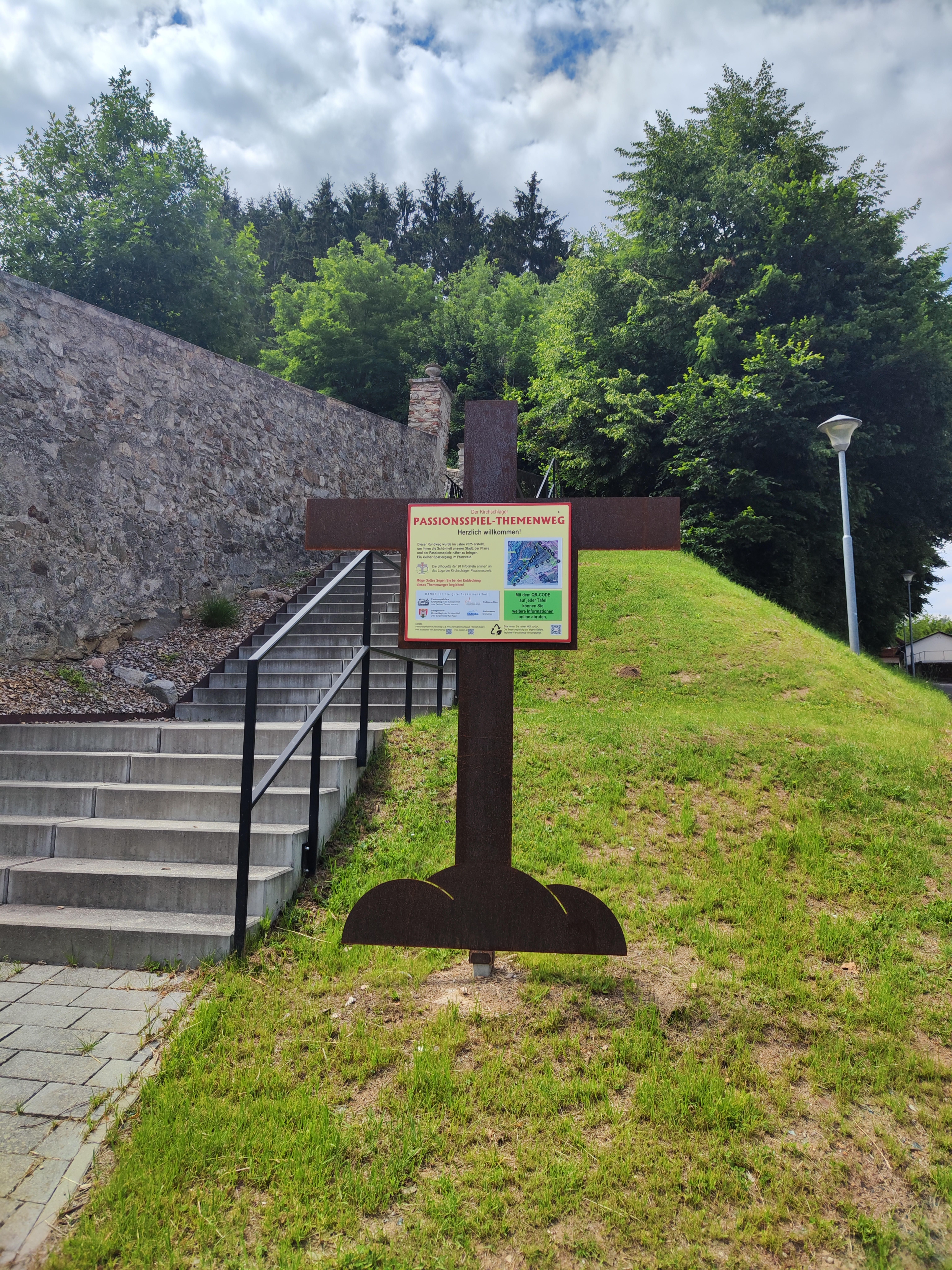 Ein Schild in Form eines Kreuzes markiert den Passionsspiel-Themenweg in Kirchschlag. Es steht auf einer Wiese neben einer Treppe und einer Steinmauer.
