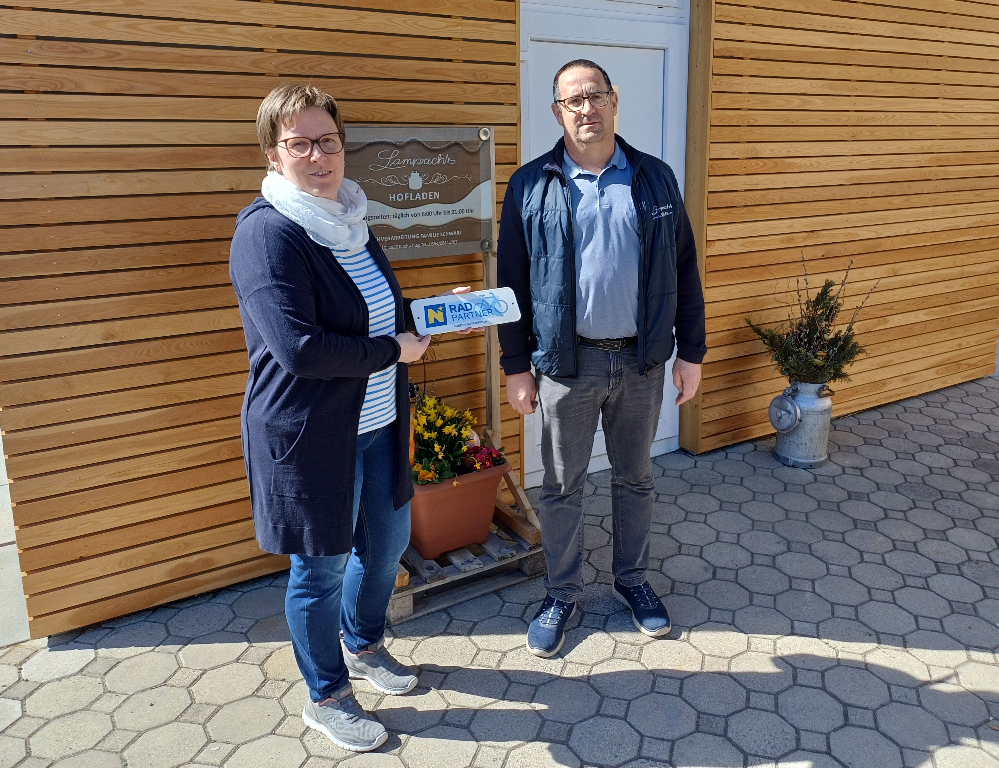Zwei Personen stehen vor einem Schild "Lamprechts Hofladen". Die Frau hält ein "Radpartner Niederösterreich" Schild in den Händen.
