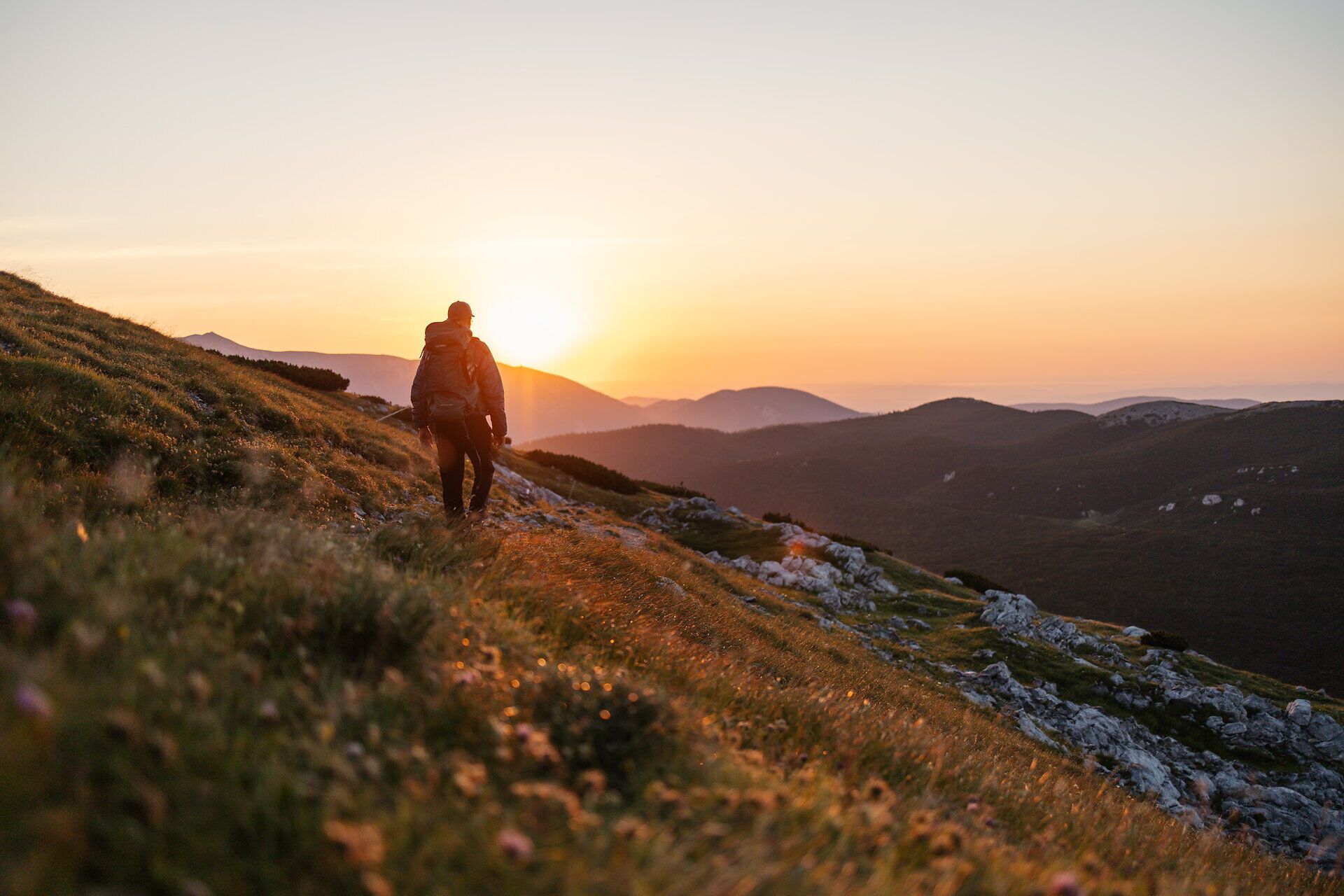 Ein Wanderer genießt den atemberaubenden Sonnenuntergang über den sanften Hügeln, während die letzten Strahlen des Tages die Landschaft in warmes Licht tauchen. Die frische Bergluft und die blühenden Wiesen schaffen eine harmonische Atmosphäre, die zum Verweilen einlädt.