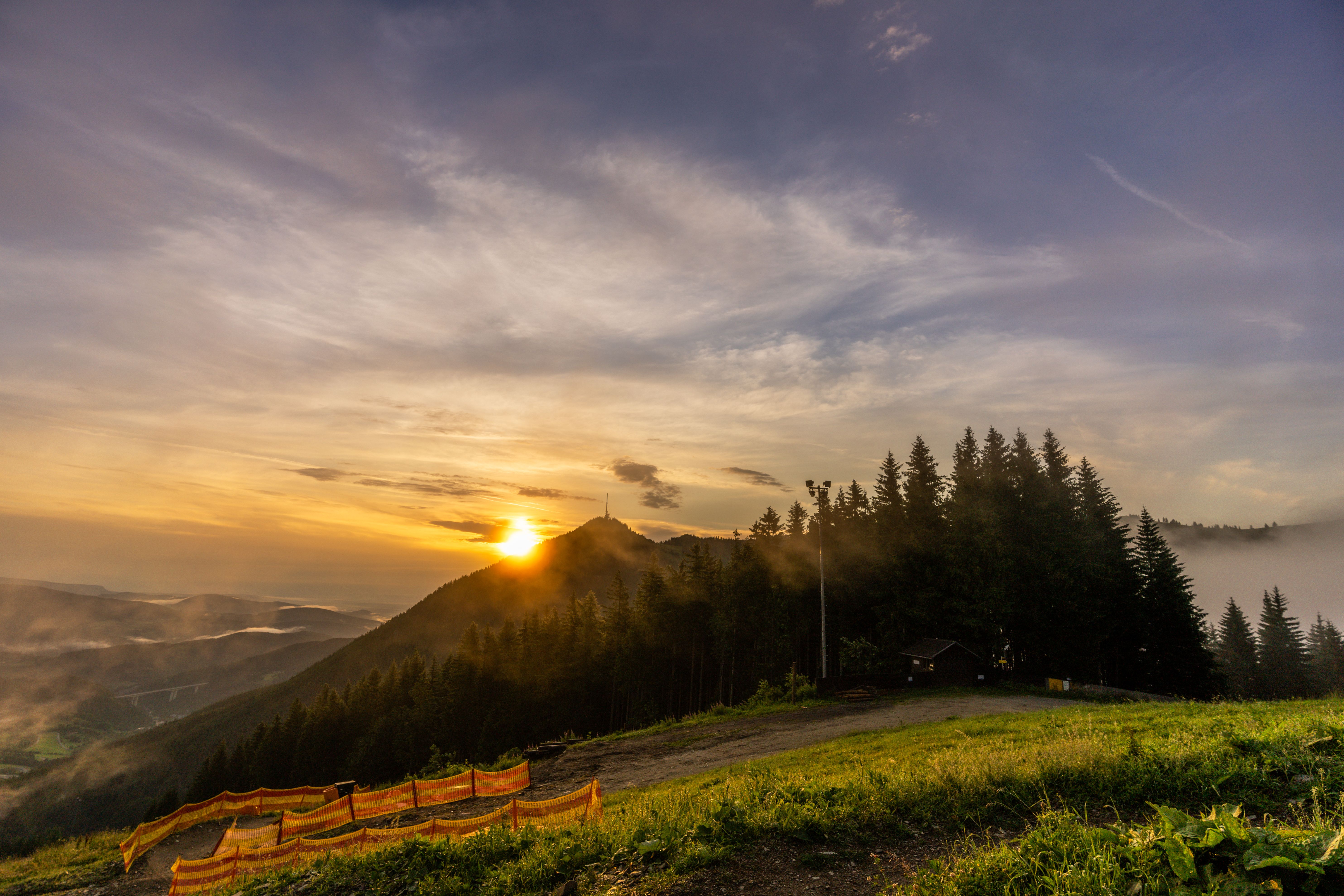 Zwei Wanderer gehen auf einem Weg bergauf, vorbei an einem Aussichtsturm und einem Gebäude, umgeben von Bäumen und einem klaren Himmel.