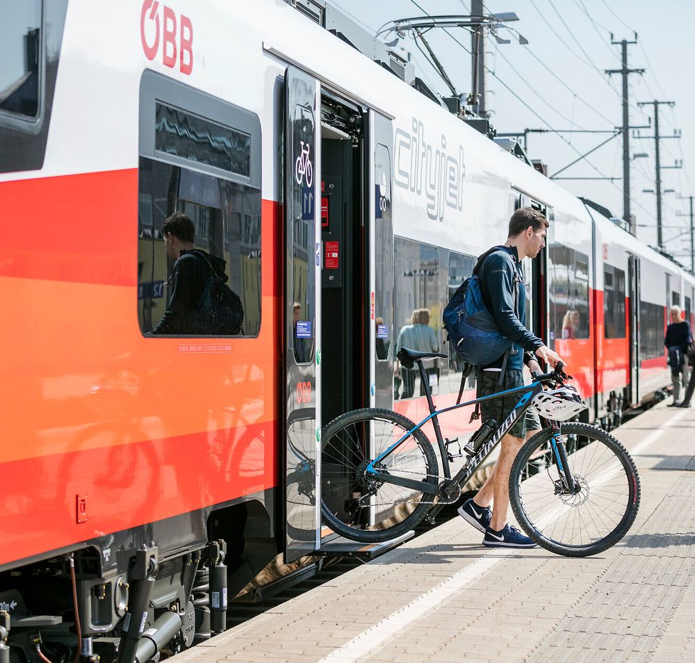 Die Sonne strahlt über den Bahnhof, während Reisende mit ihren Fahrrädern in den Zug einsteigen. Die frische Luft und die Vorfreude auf neue Abenteuer in der Natur sind spürbar. Ein perfekter Start für eine unvergessliche Reise in die Berge.