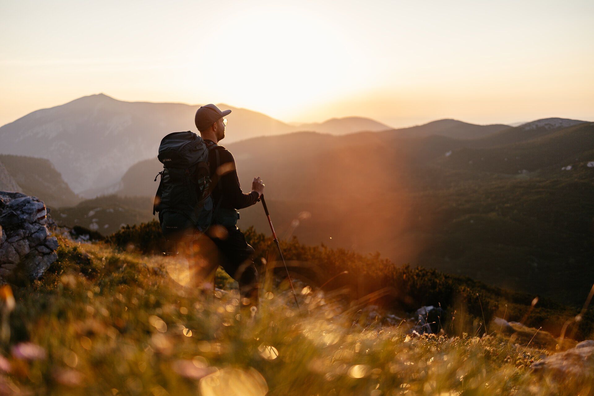 Ein Wanderer genießt den atemberaubenden Sonnenuntergang über den majestätischen Bergen, während die sanften Hügel in warmen Goldtönen erstrahlen. Die frische Bergluft und die unberührte Natur schaffen eine perfekte Kulisse für unvergessliche Erlebnisse in den Wiener Alpen.