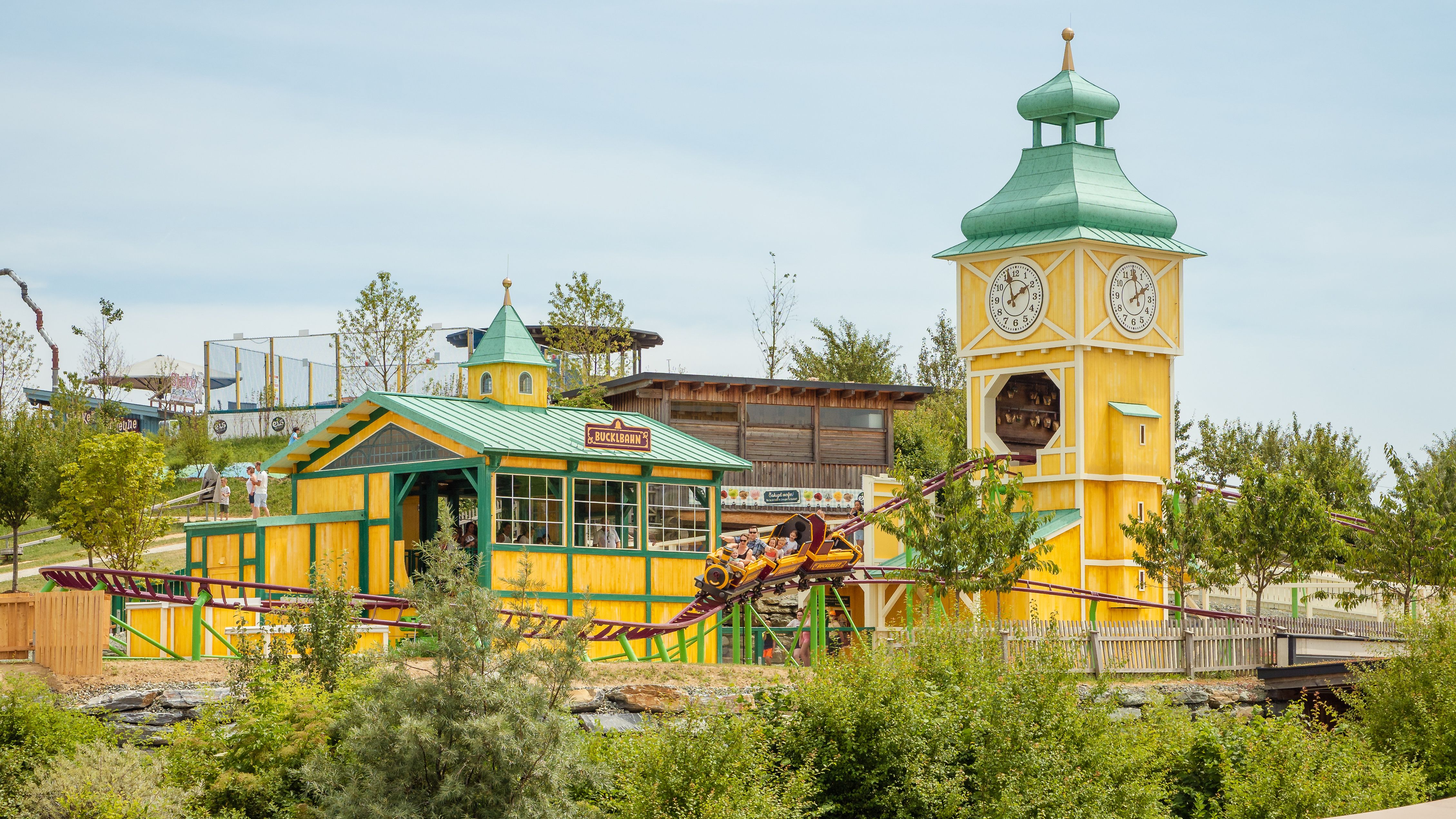 Die Bucklbahn, eine Achterbahn, auf roten geschwungenen Schienen und mit gelb-grünem Gebäude und Uhrturm, rundherum wachsen grüne Sträucher