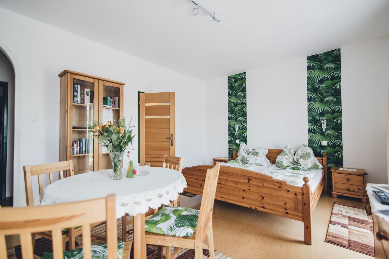 Ein gemütliches Doppelzimmer mit Holzmöbeln, einem Tisch mit Blumen, einem Bett mit Pflanzenmuster und einem Bücherregal.