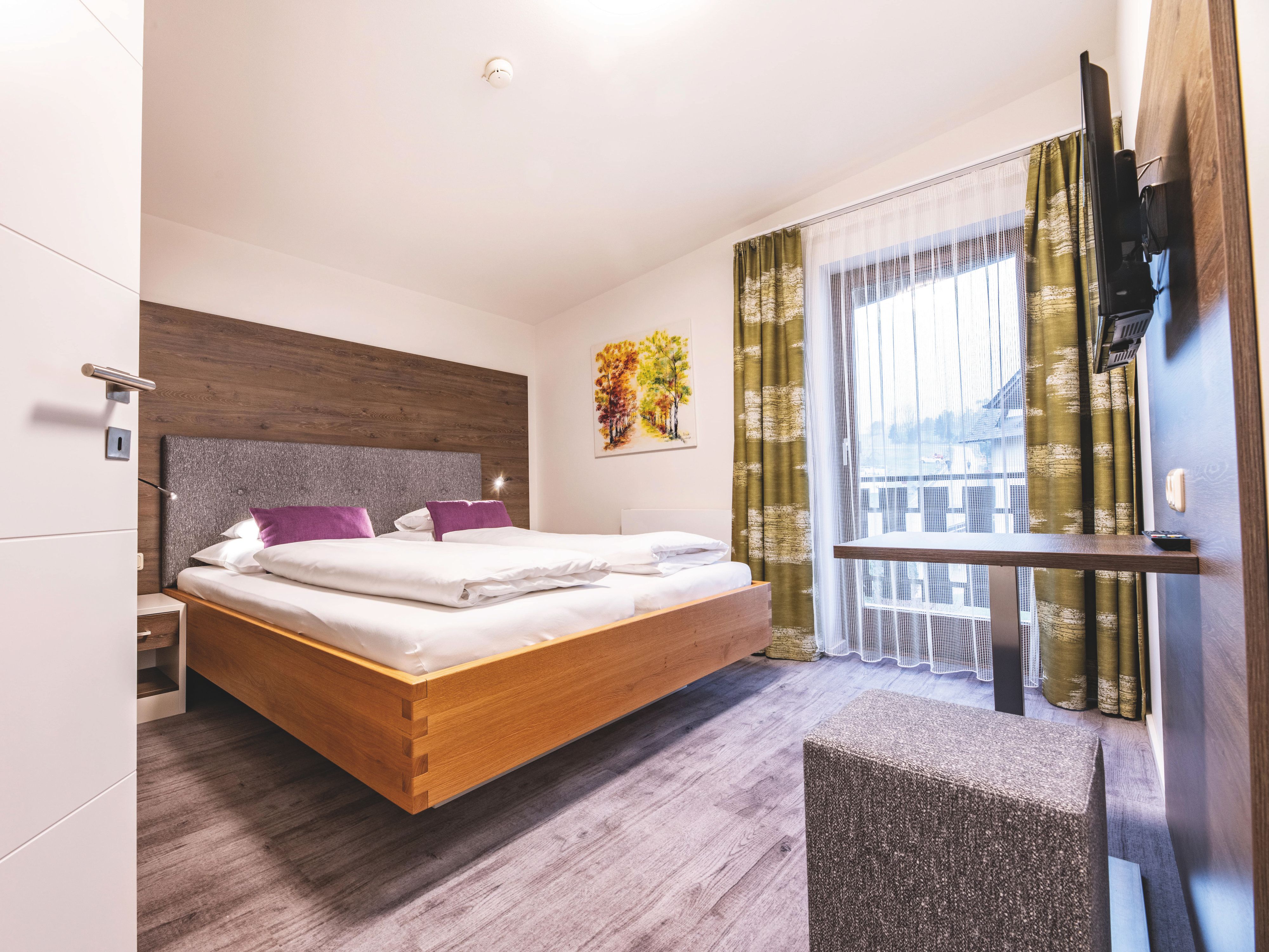 Ein modernes Hotelzimmer mit Terassentür mit Vorhängen, Fernseher und Tisch mit Hocker.