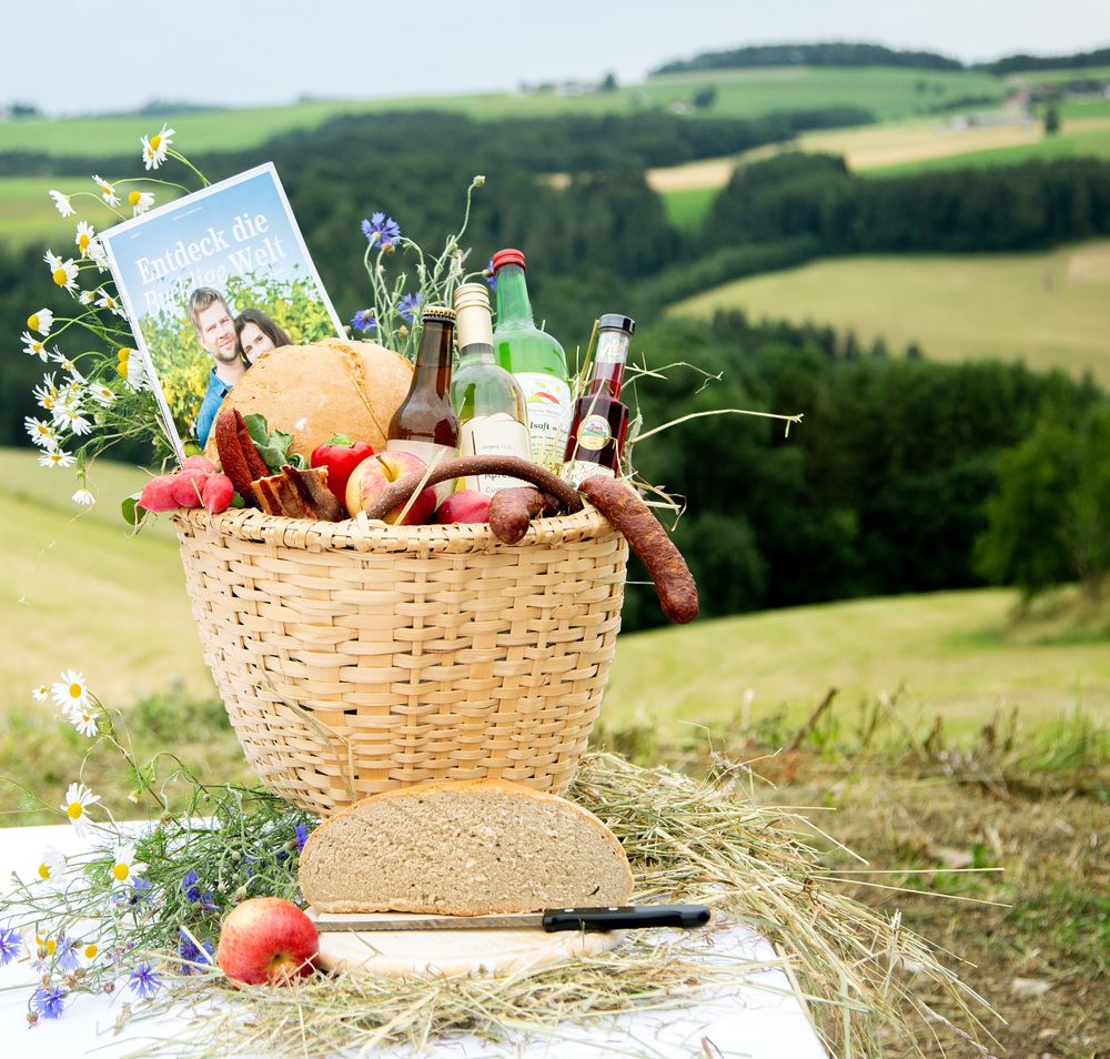 Ein Bucklkorb mit regionalen Lebensmitteln, davor Brot und ein Apfel, im Hintergrund grüne Landschaft.