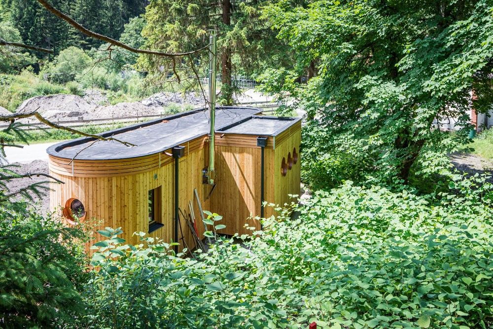 Ein kleiner, hölzerner Wohnwagen steht inmitten von Bäumen und Büschen.