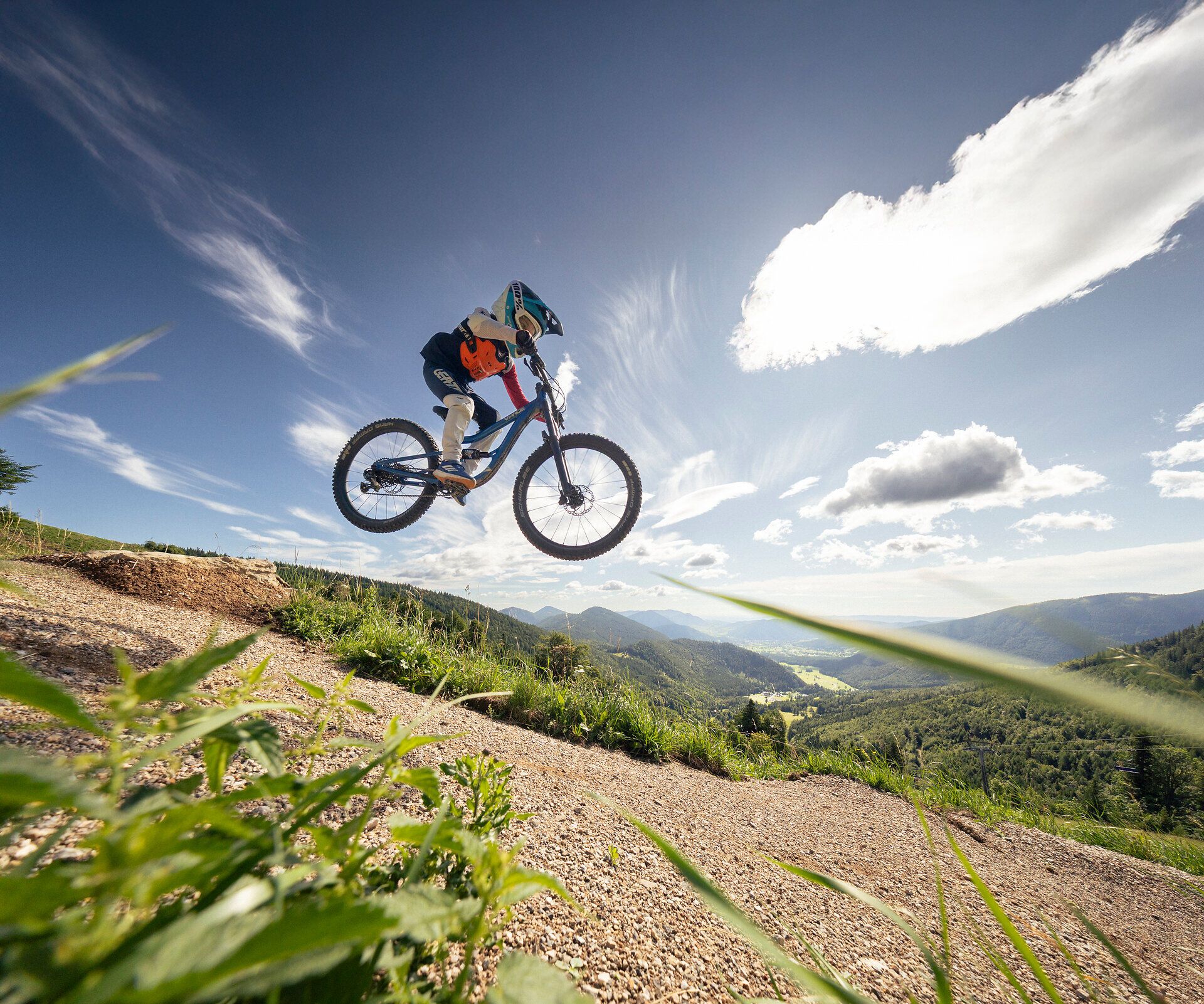 Mountainbiken im Alpenbikepark Puchberg am Schneeberg in Niederösterreich: Kind springt mit dem Mountainbike über einen Sprung auf einem alpinen Trail, mit Blick über die Wiener Alpen.