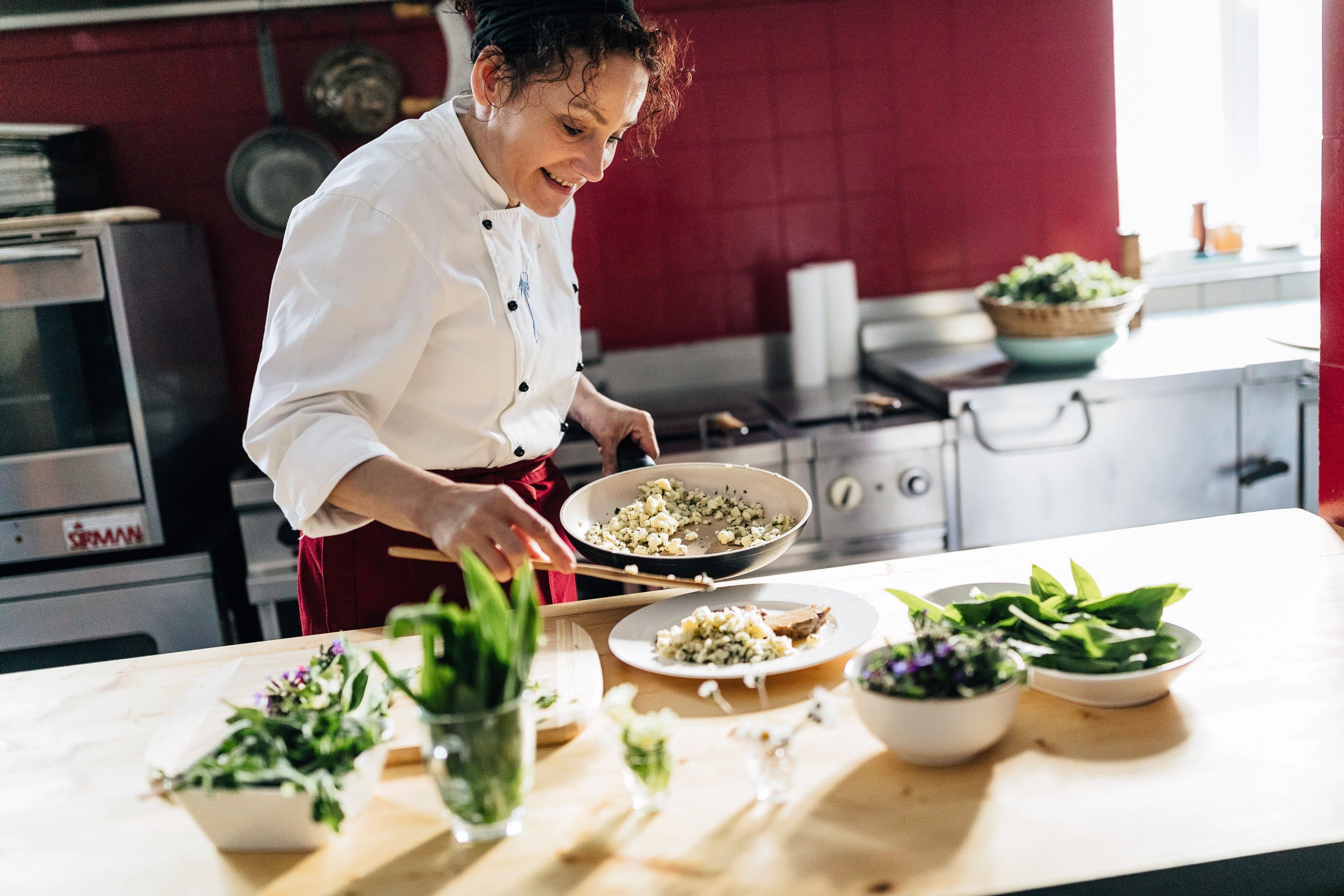 Eine Köchin hält eine Pfanne und richtet ein Teller mit einem Gericht an.